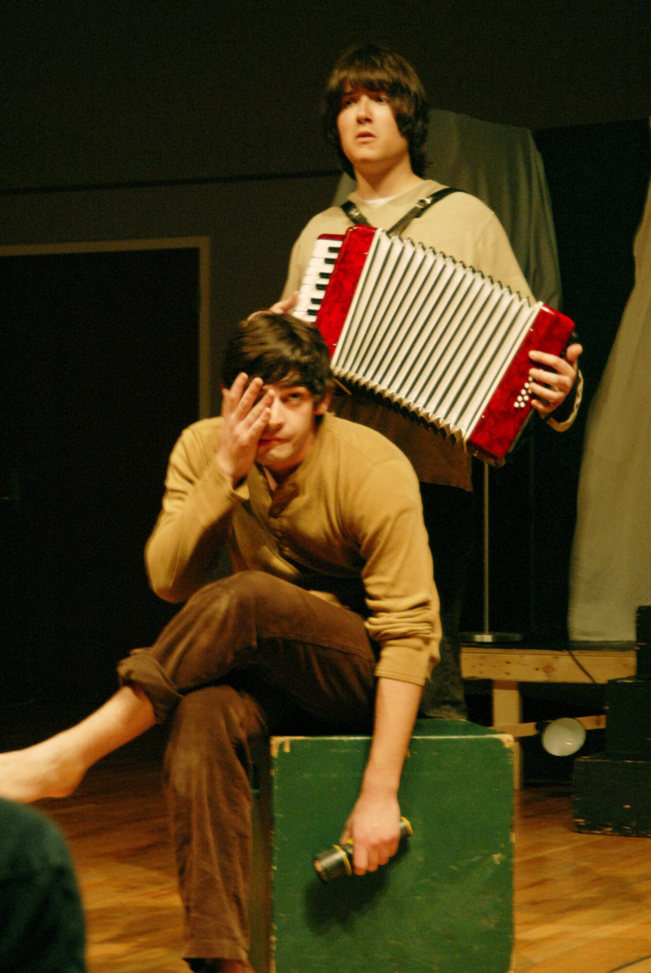 One student seated on a green block, another stands behind him, playing an accordian.