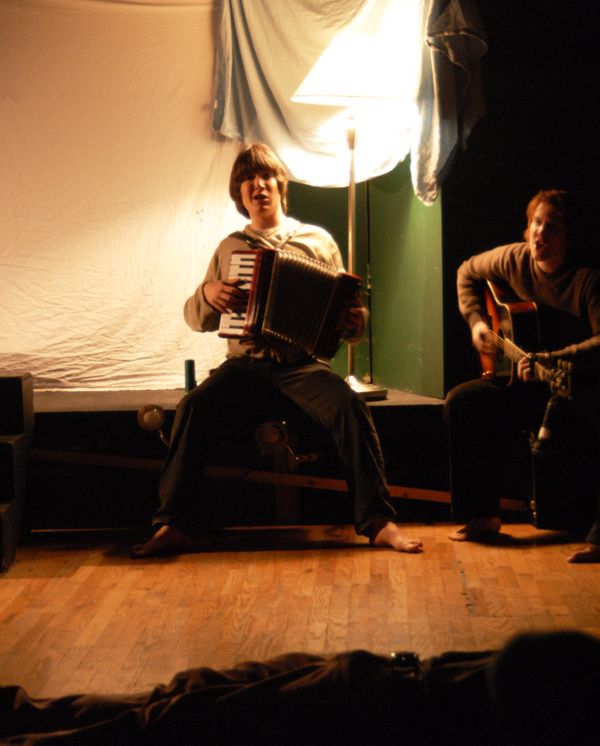 One student plays an accordion in front of a white sheet, another sits next to him playing guitar.