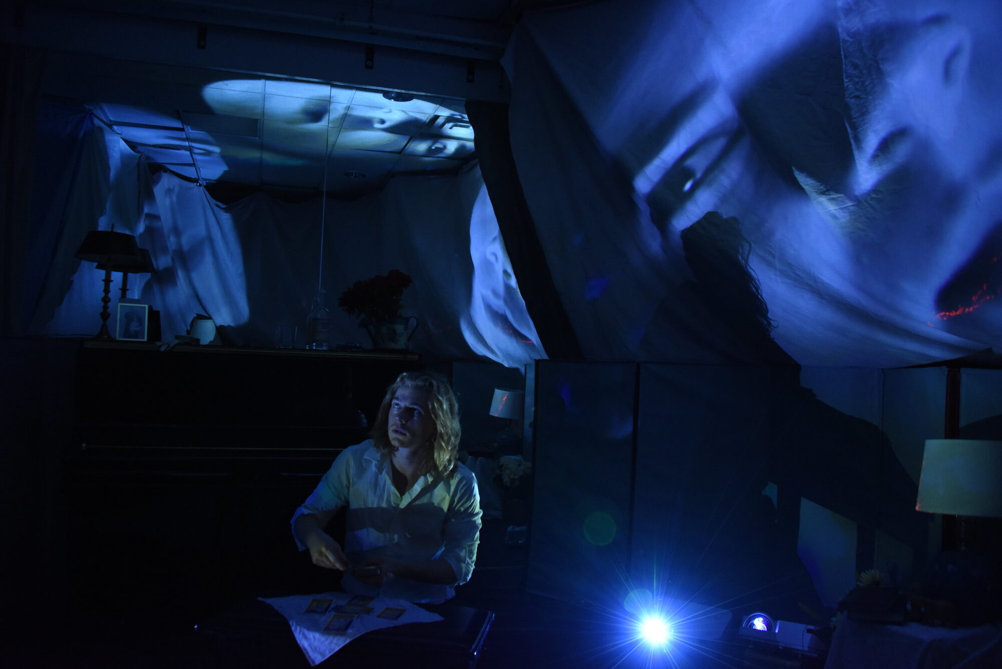 An actor sits in a small dark space with projections of faces on the surrounding walls.