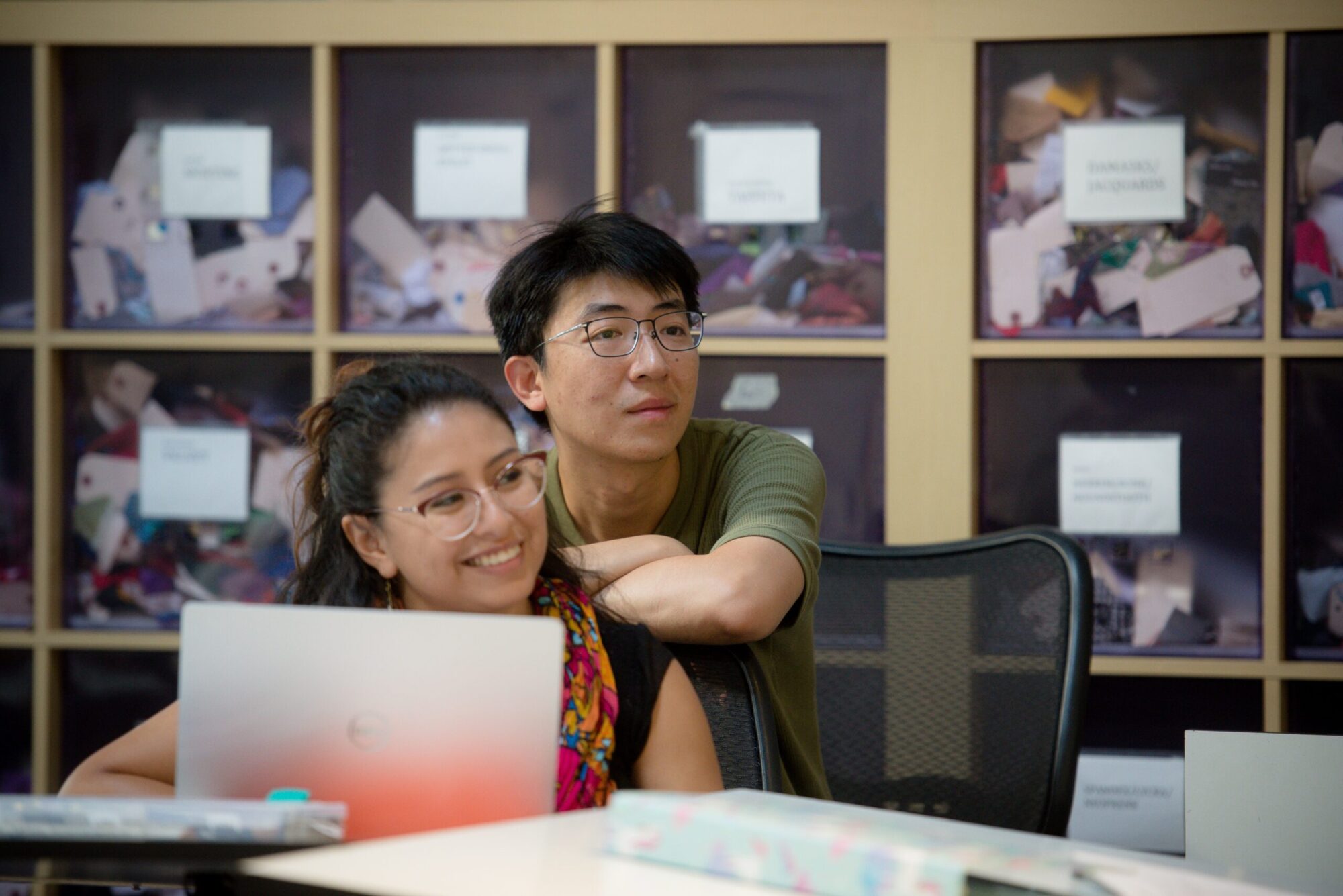 Two students sit at a design table in a costume studio, with a laptop open, leaning in and smiling.