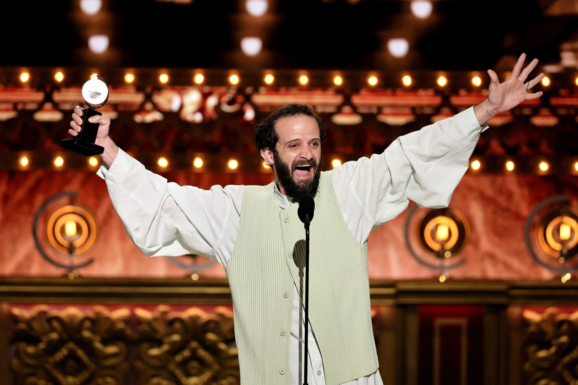 Actor and alumnus Will Brill stands on stage accepting his Tony Award, wearing a light green tunic and stretching his arms out wide.