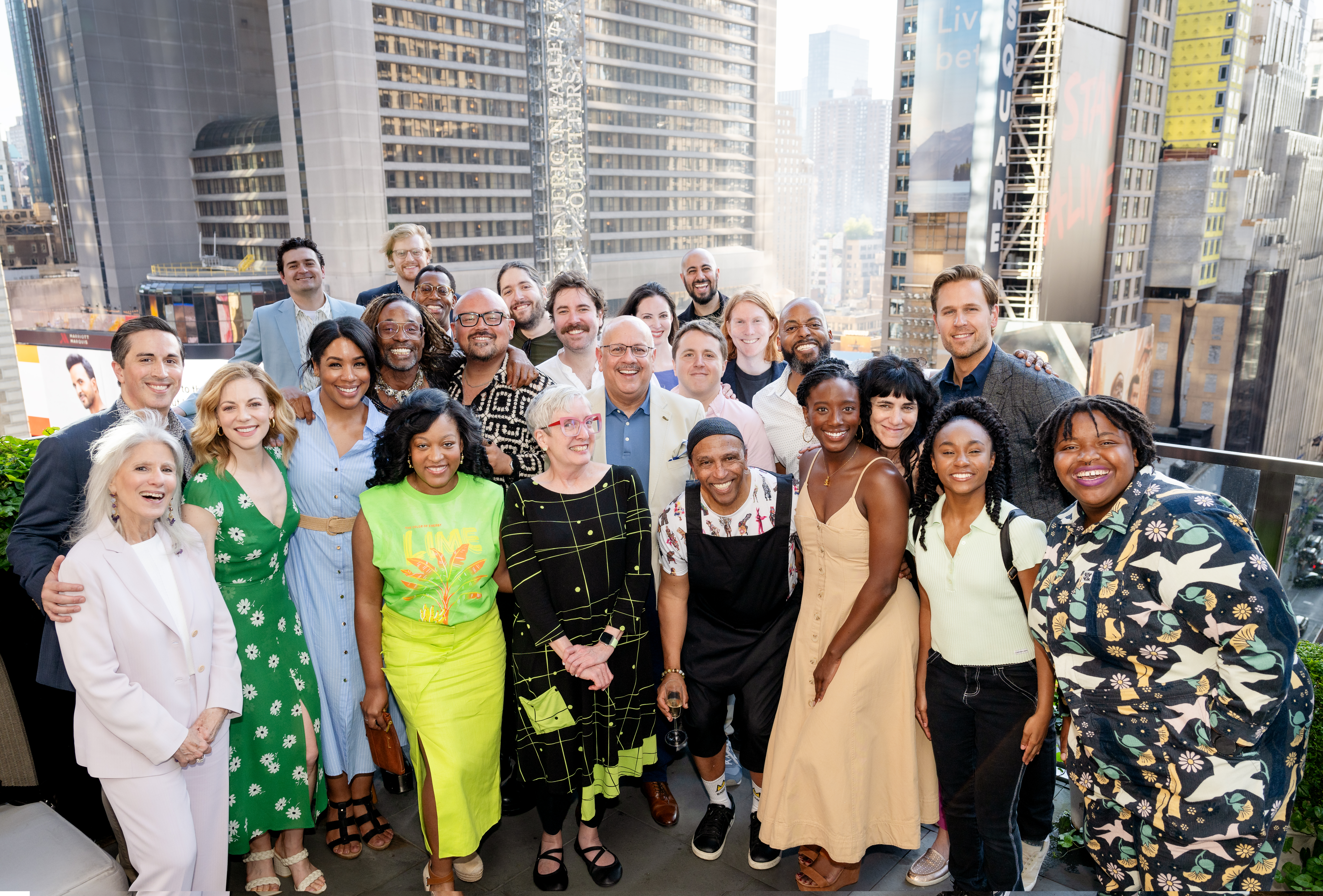 A large group of School of Drama alumni pose on a balcony in NYC, smiling at the camera, which shoot them from above, the city scape in the background.