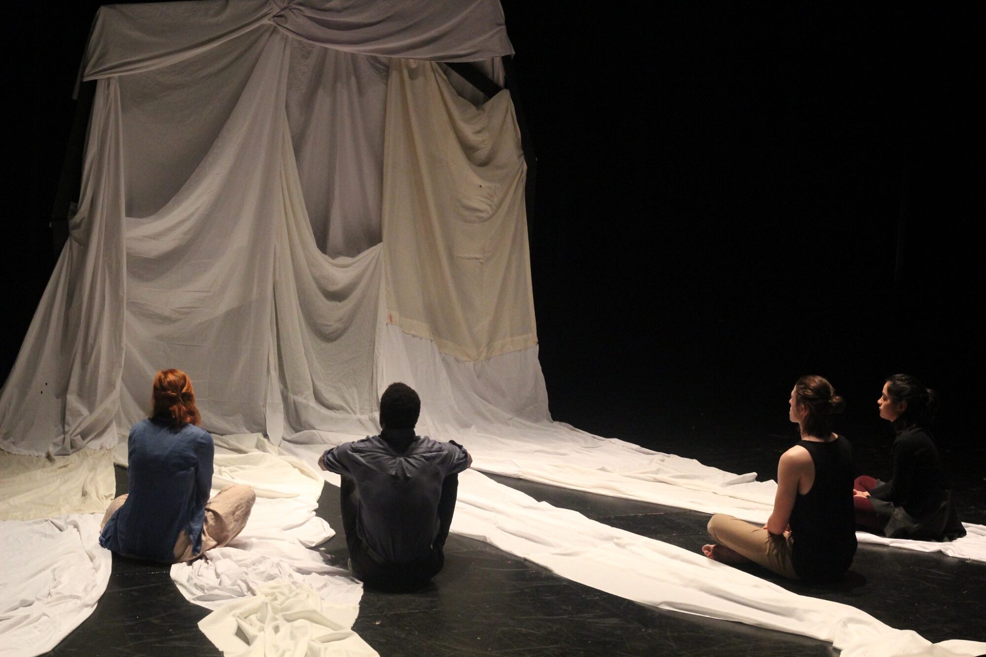 Large white sheets drape the walls and floor of a black box theater, with students seated on the ground facing the sheets.