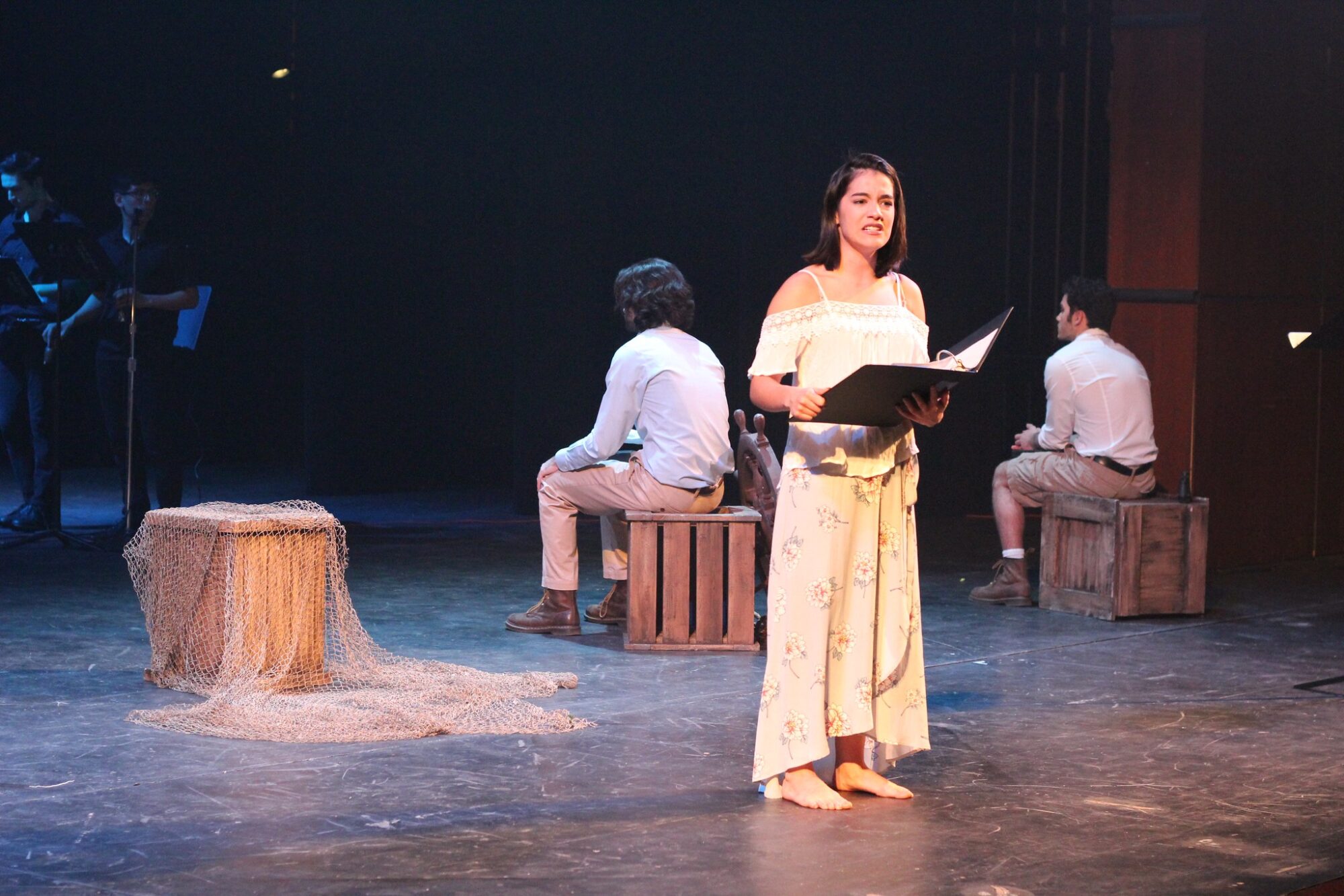 Three students wearing all white are on stage, two sit facing away from the audience, the third holds a script and looks out into the audience.