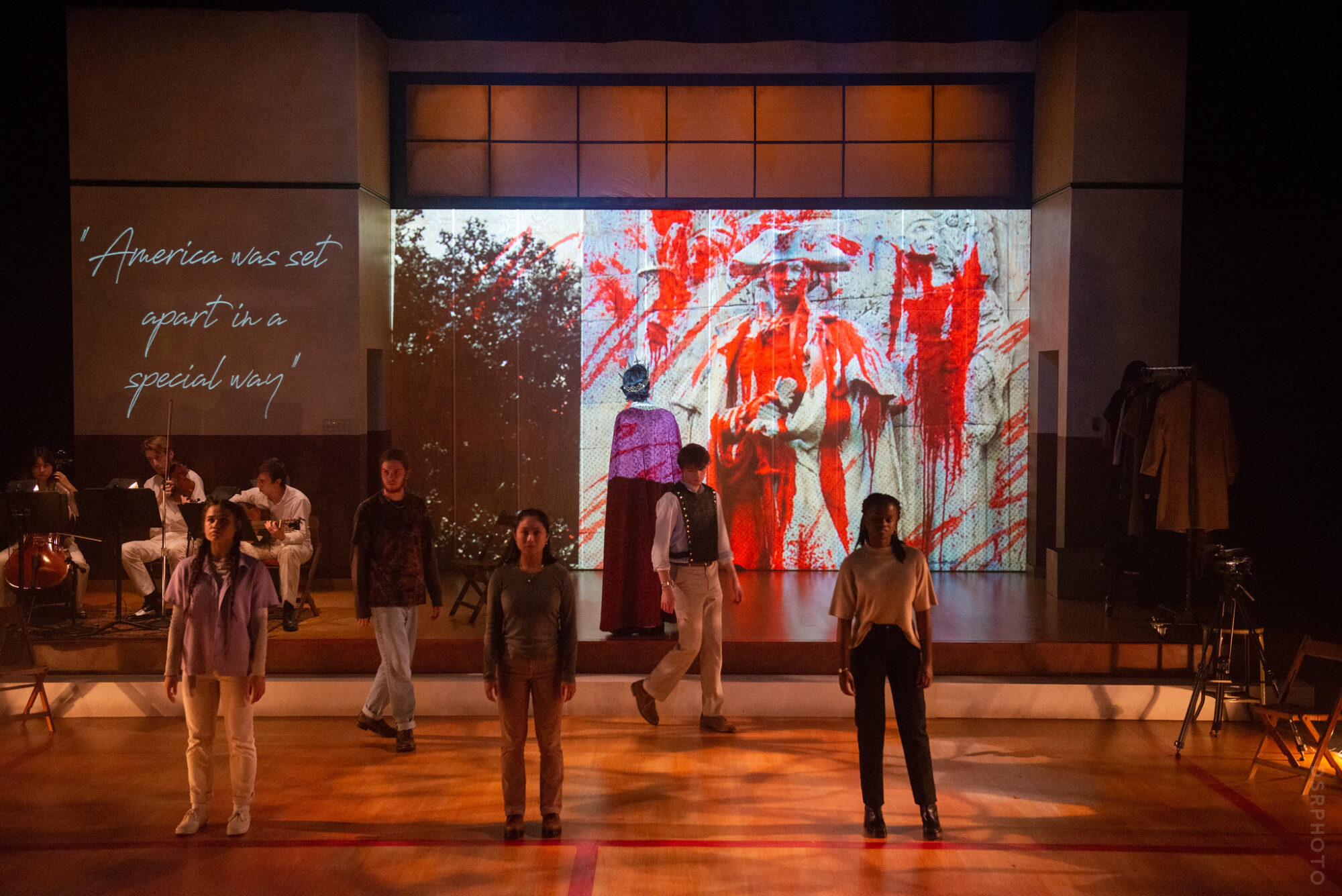 A group of actors on stage, behind them images and text are projected onto the back wall.