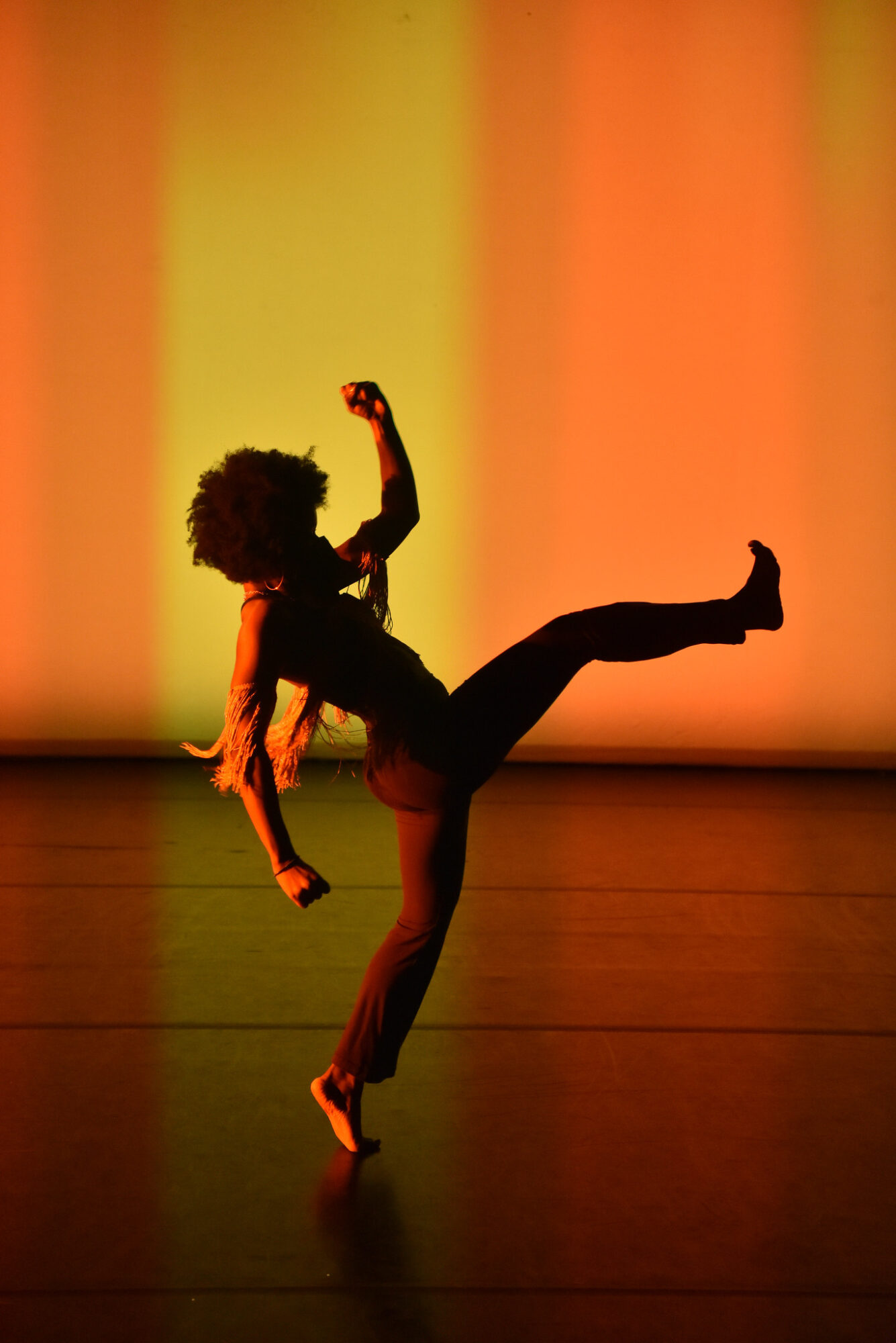 A dancer in silhouette kicking one leg up, with arms in alternate positions, against a bright orange and yellow lit backdrop.