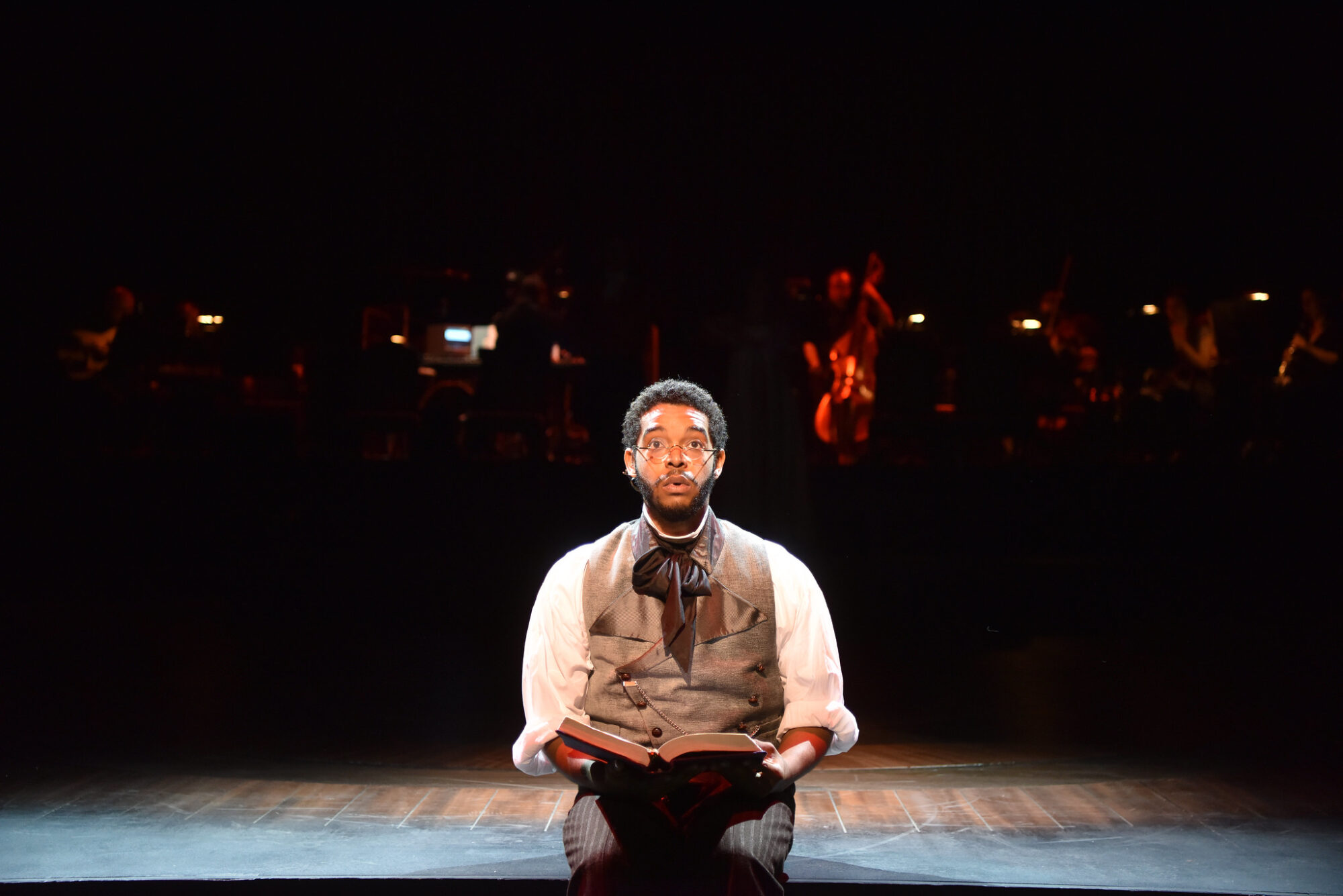 A student actor in costume sits on the edge of a stage with an open book in his lap, looking out into the audience.