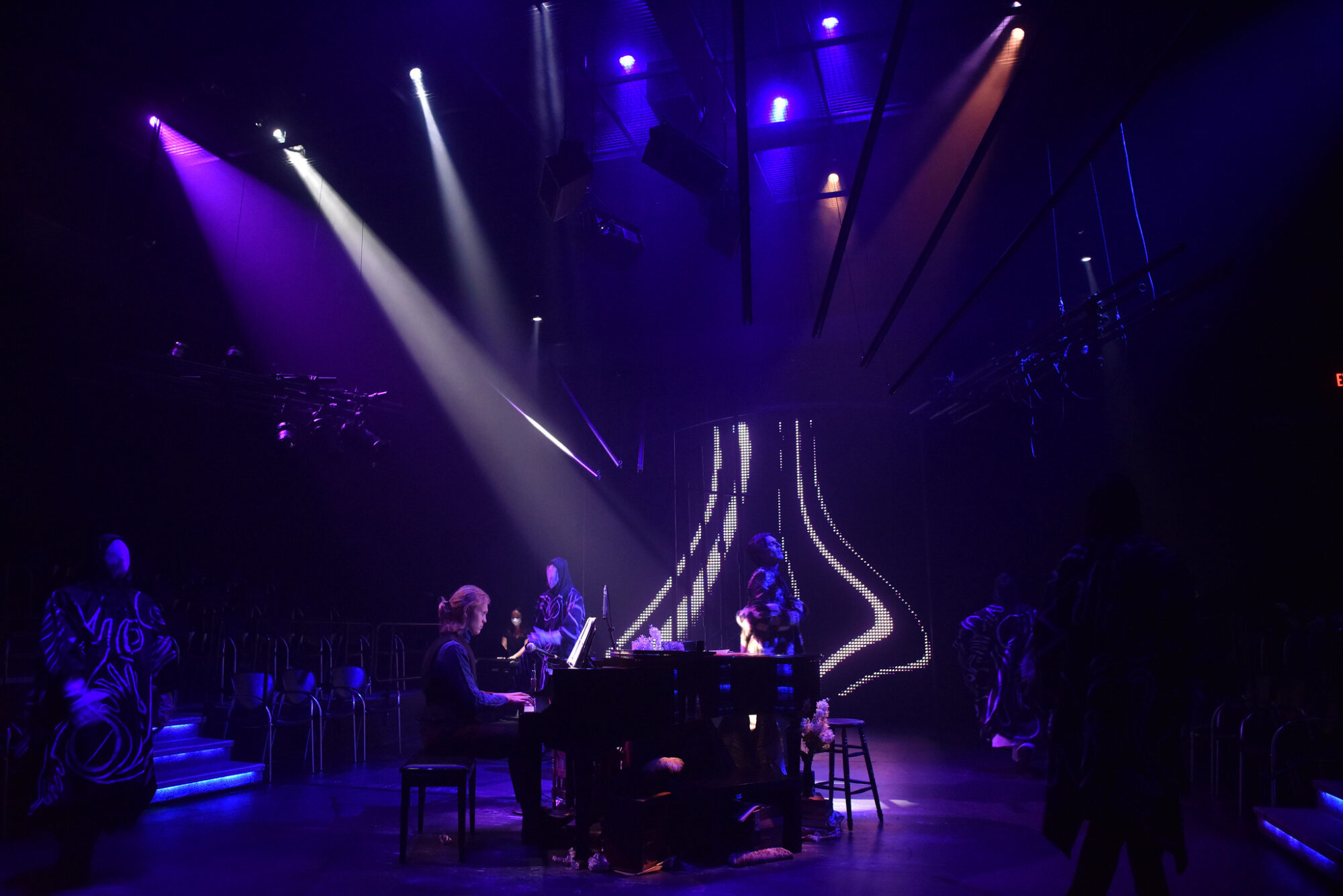 Dramatic purple and blue stage lighting shine down onto a performer sitting at a piano in a production of “Preludes” at the School of Drama.