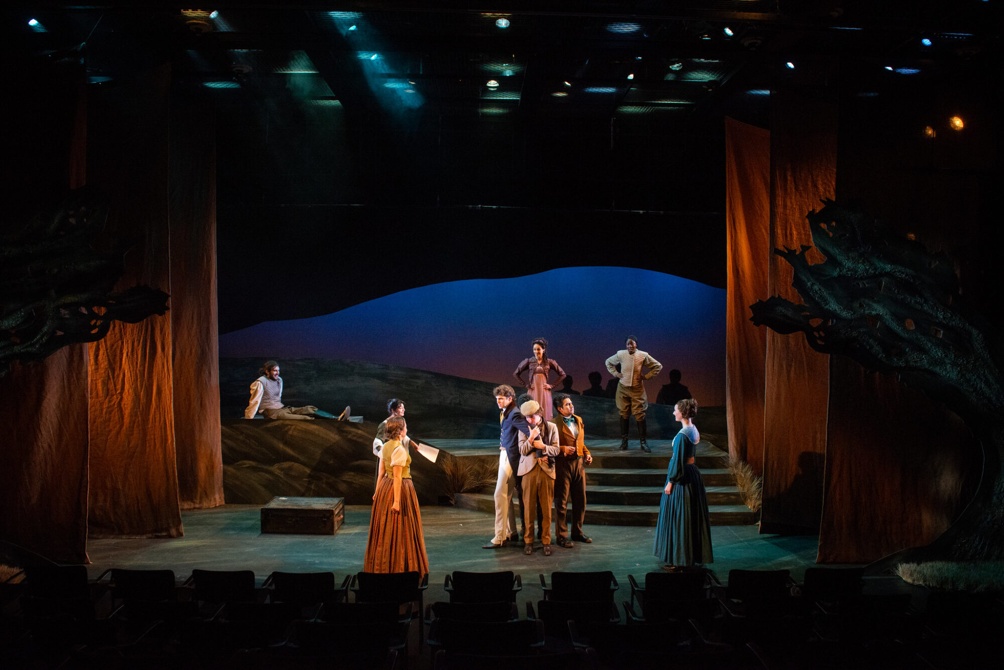 Nine actors in period costumes on a set that contains stairs leading to a second level, with rolling hills in the background and curtains hanging on either side; a scene from “You on the Moors Now.”