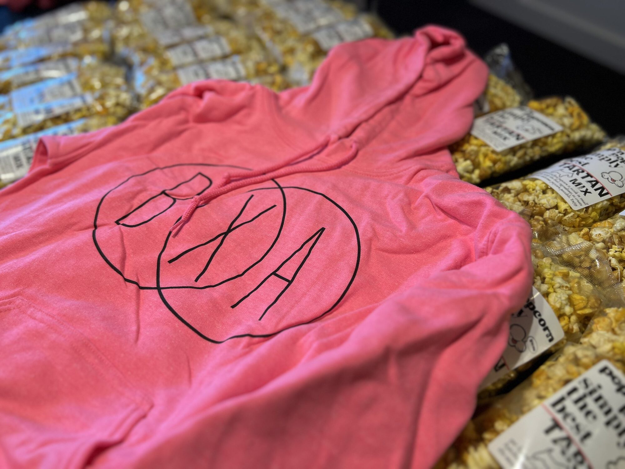 A pink hooded sweatshirt with two concentric circles and the letters BXA in black ink lays on top of a table full of packages of caramel popcorn.