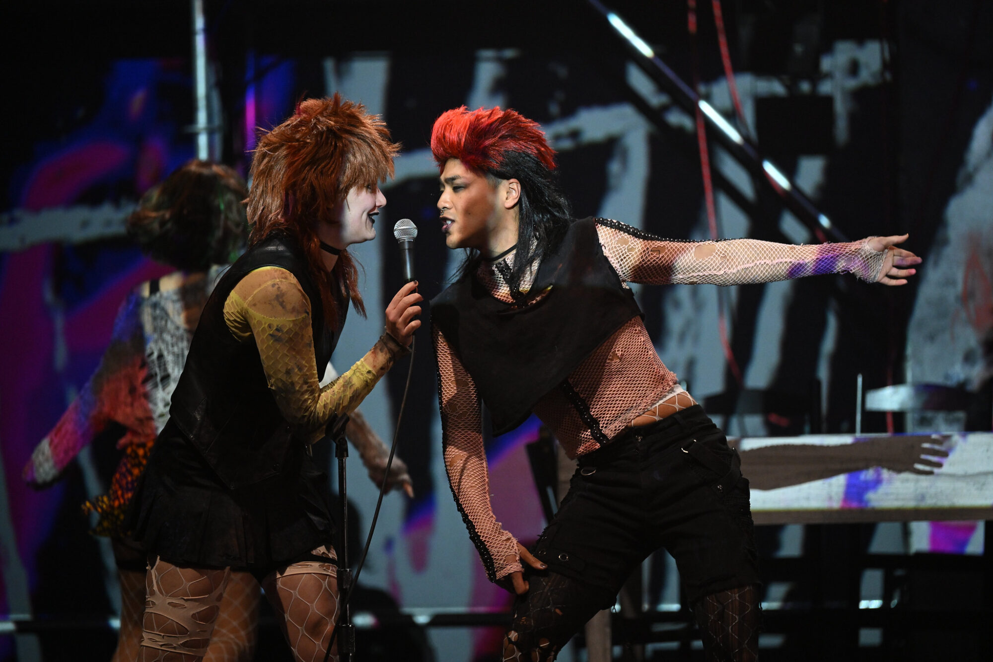 In a scene from “I Think I’m Starting to Want to Be a Mother,” two students on stage in punk rock costumes and wigs sing into a standing microphone.