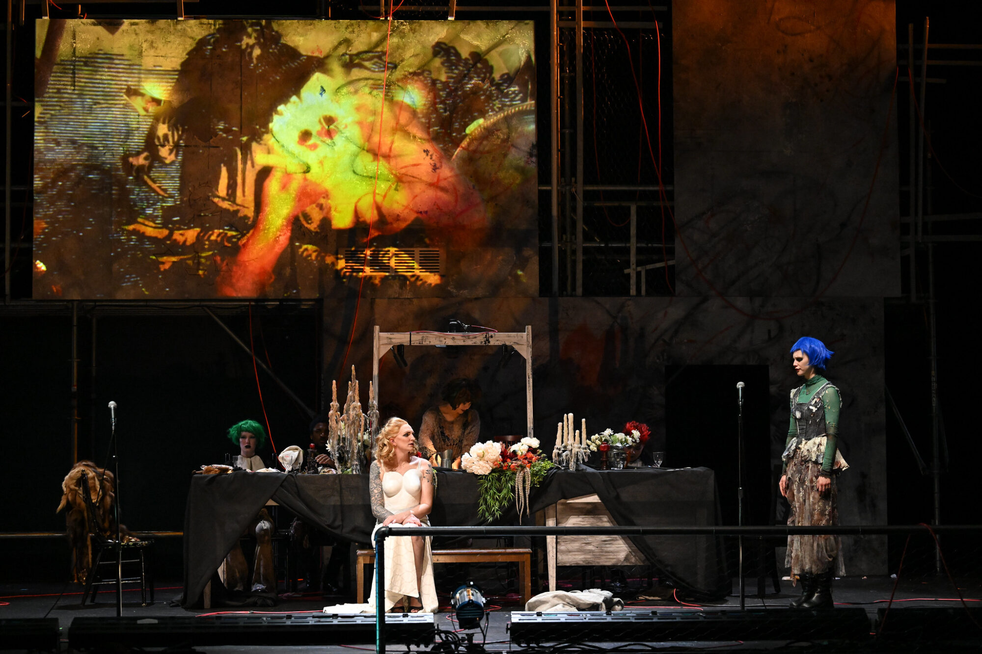 Actors sitting at a decadently decorated table, wearing pop punk costumes and wigs, with a large projected image on the wall behind them.