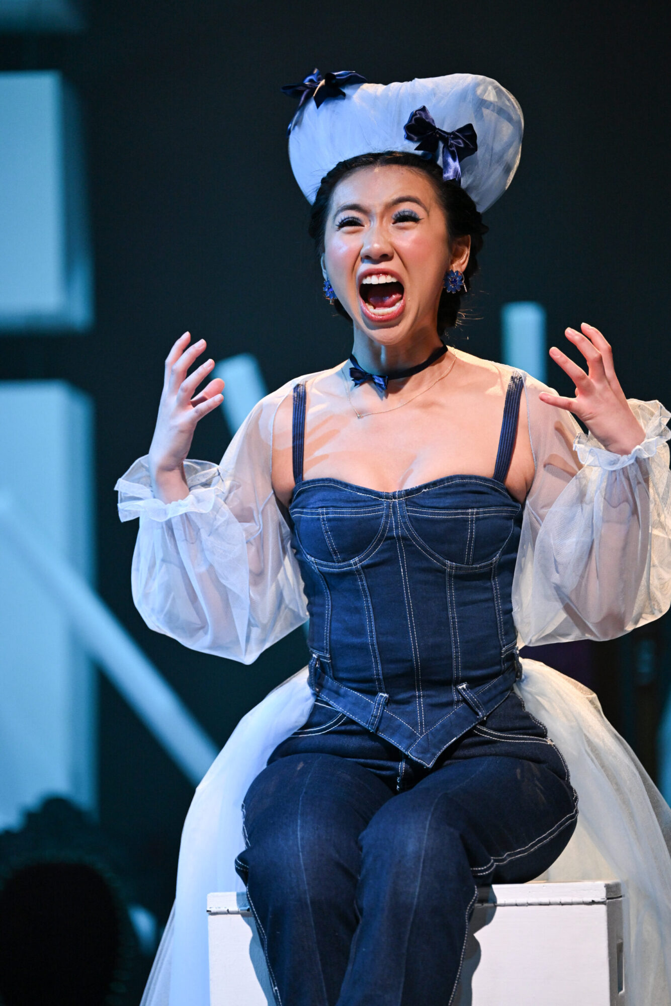 Student Helena Chuang, wearing a denim jumpsuit and a period costume headpiece, sits on top of a set piece, screaming and gesturing with her hands in a scene from “Lear.”