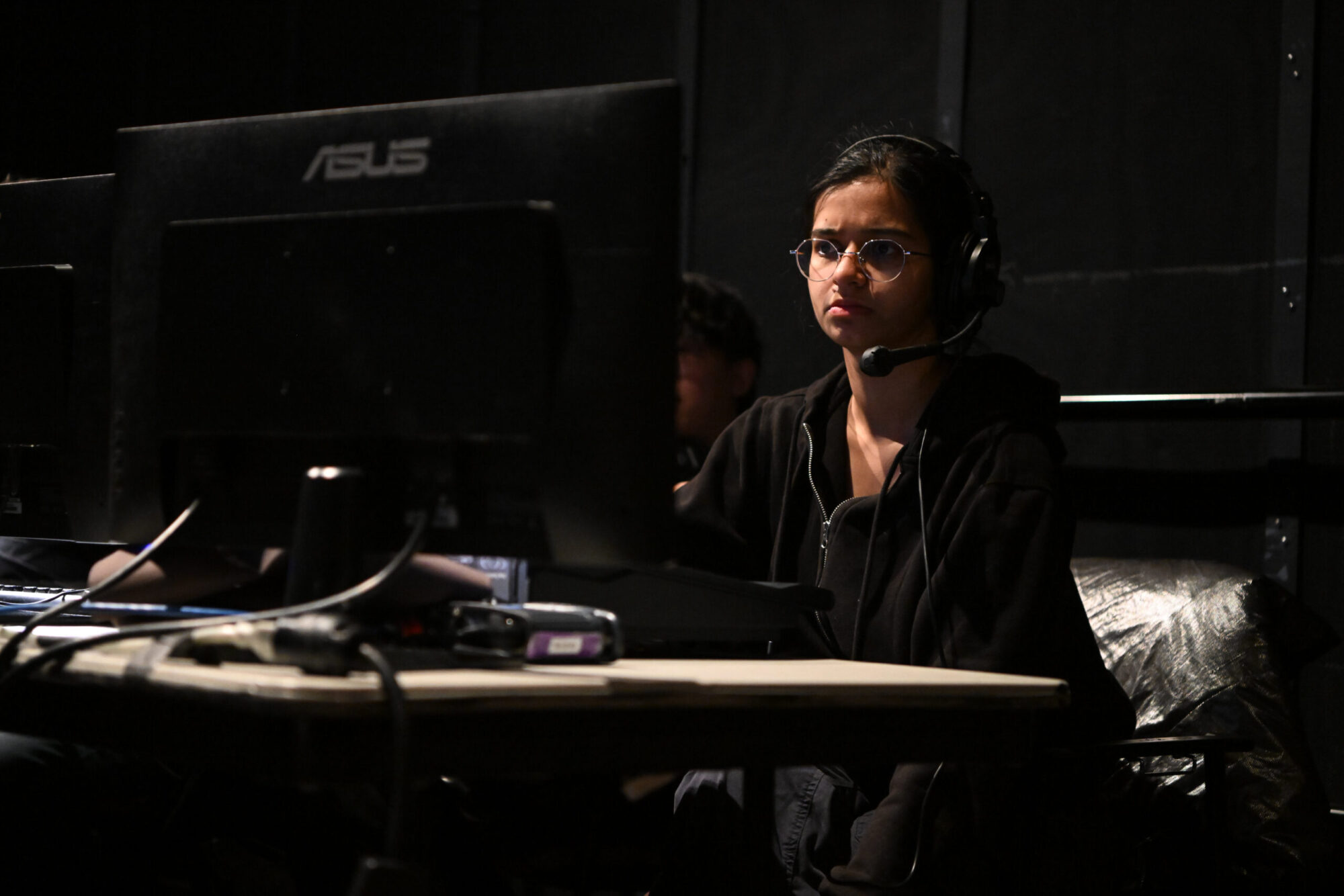 A lighting design student wearing all black and a headset sits at a table with a large computer monitor in a dimly lit theater space.