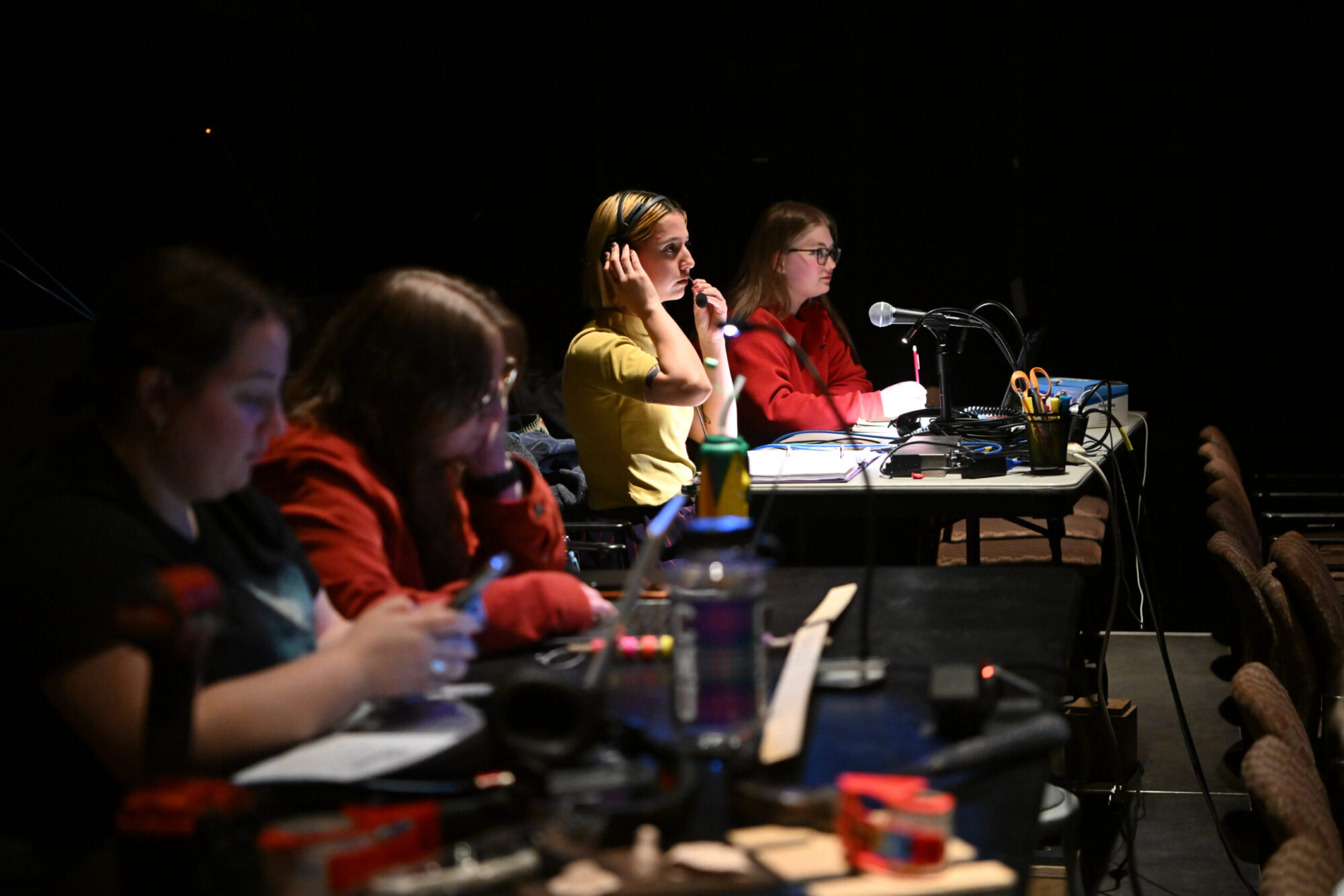 Four students sit at tech tables in rehearsal for a play; the student in focus speaks into a headset.