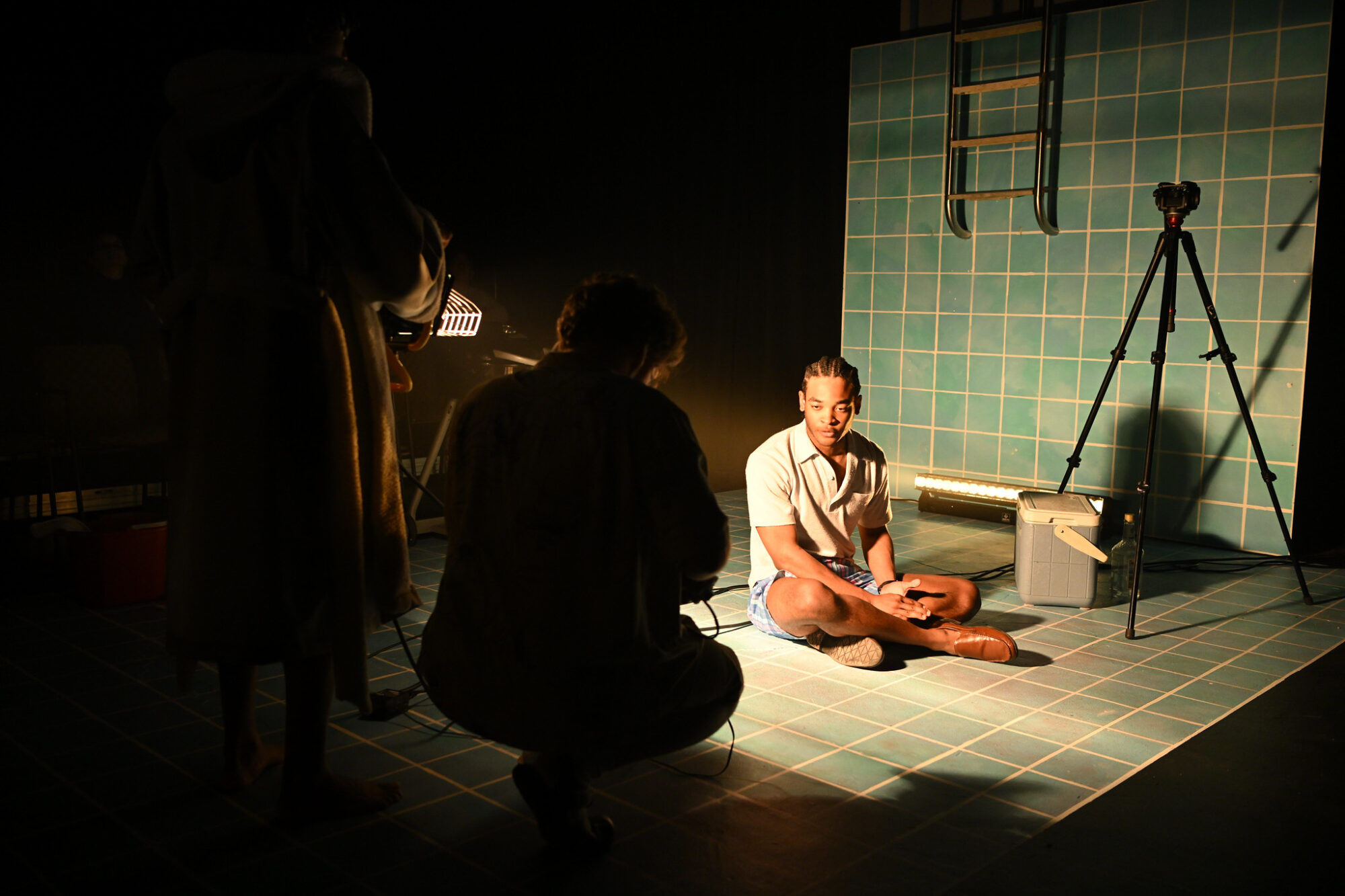 Student Carlwell Redmon sits on a set that looks like the bottom of a swimming pool in a spotlight, being filmed by figures in silhouette in a scene from “Penelope.”