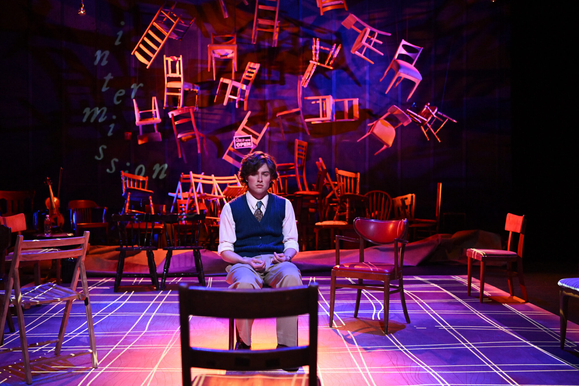 A student actor sits on a chair on stage, with many chairs suspended and hanging behind him, in a scene from "Lonely Planet."