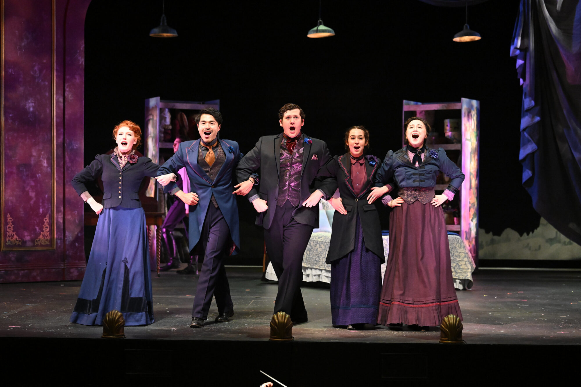 Five students stand on stage in period costumes with arms linked, singing during a performance of "A Little Night Music."