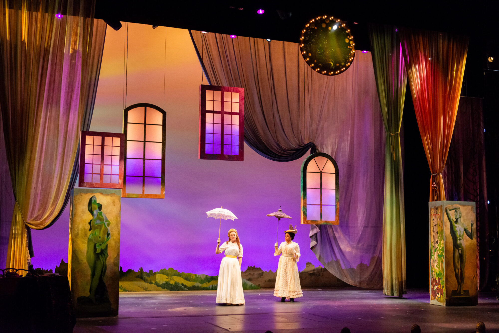 A stage beautifully lit in orange and purple sunset colors, window frames suspended from the ceiling, curtains draped off to the sides, and two actors wearing white dresses and holding parasols are center stage.