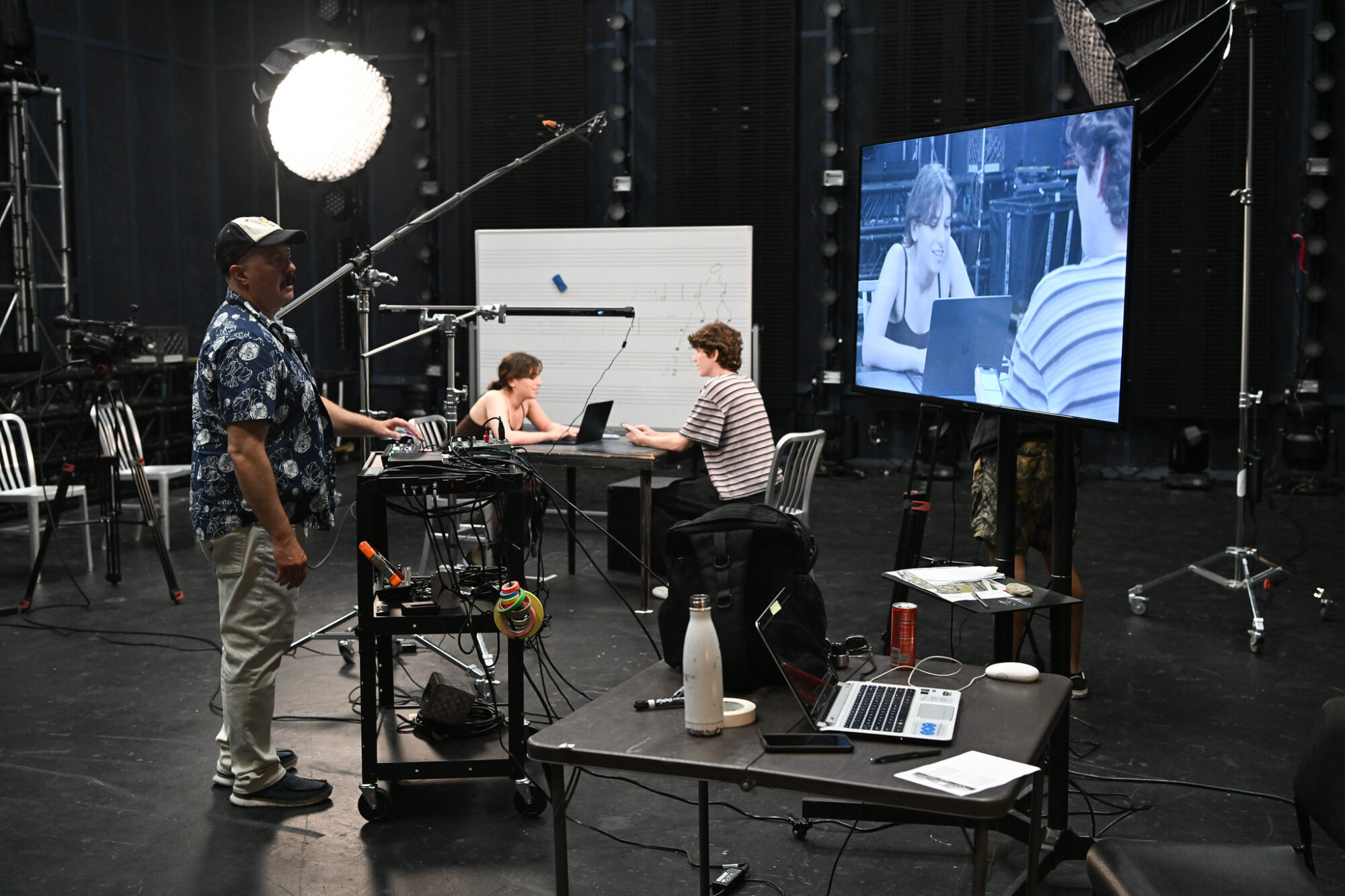 A professor and students working on a film project in the John Wells Video Studio; a large light, camera, and screen showing the students sitting at a table, while the professor watches their scene.