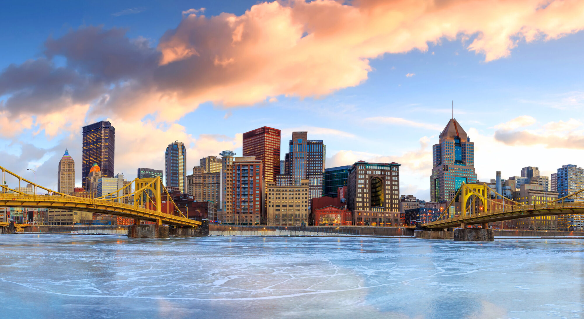 A view of Downtown Pittsburgh taken from across the Allegheny river, two yellow bridges on each side, with downtown high-rises and blue sky with large white and pink clouds overhead.