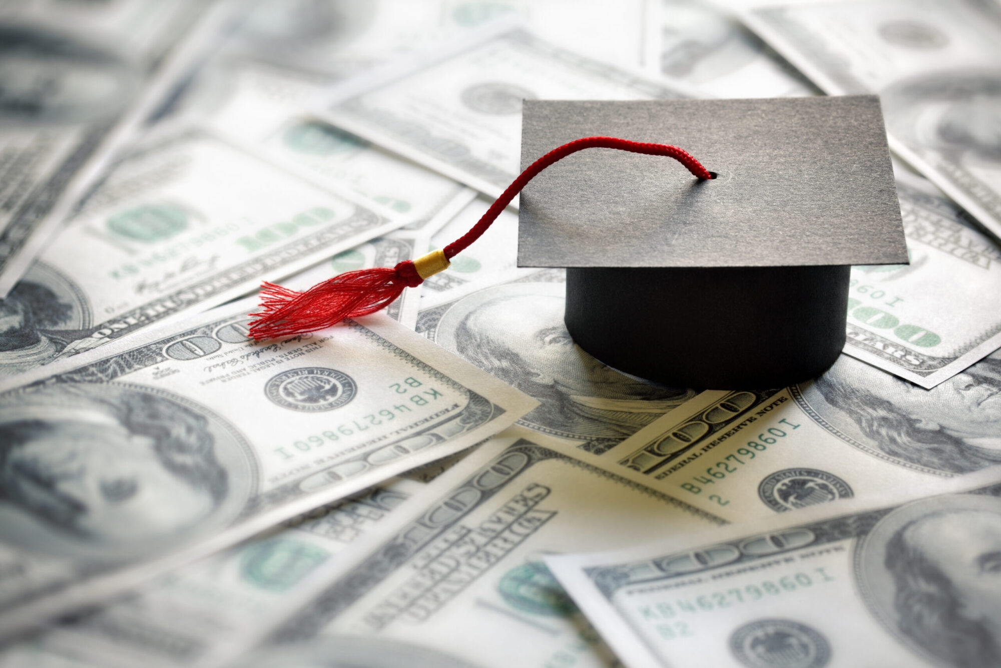 Several one hundred dollar bills are spread out with a small black graduation cap with a red tassel on top of them.