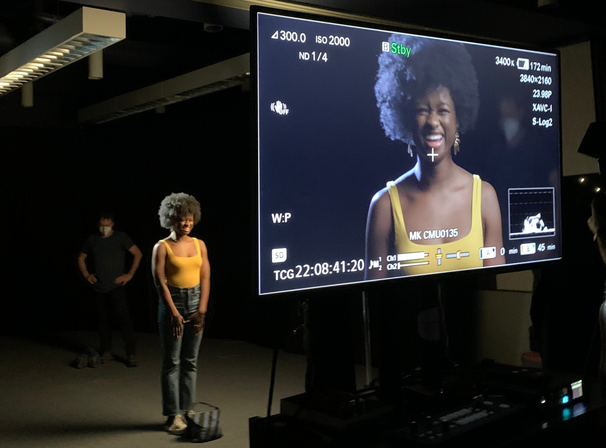 Student Ayana Cymone, wearing a yellow tank top and jeans, stands in front of a camera, her image is projected on a large screen in the foreground.