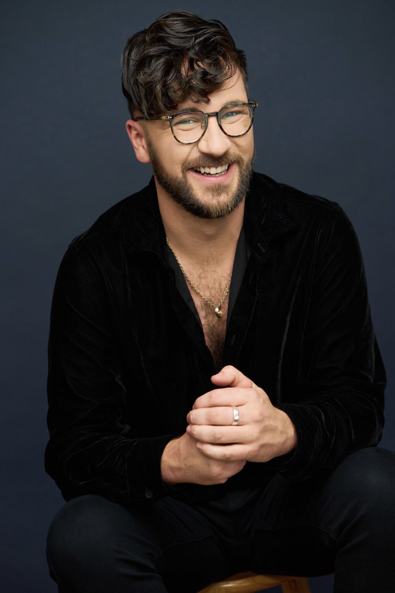 Nate Bertone, a man with brown hair, glasses, and a beard, wearing all black, looks at the camera and smiles, his hands clasped in front of him.