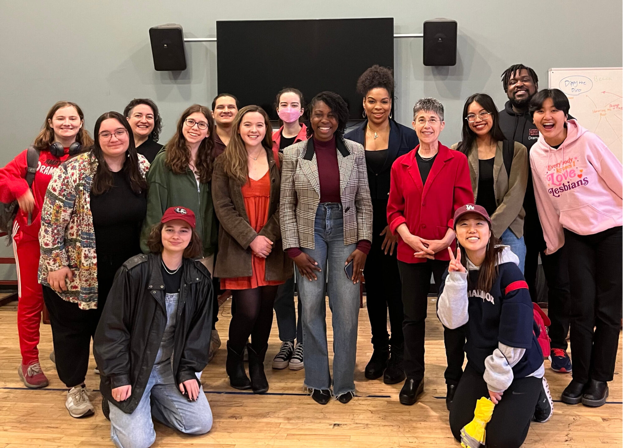 A group of students in a classroom stand and kneel, posing for a photo with alumna Alesia Etinoff.