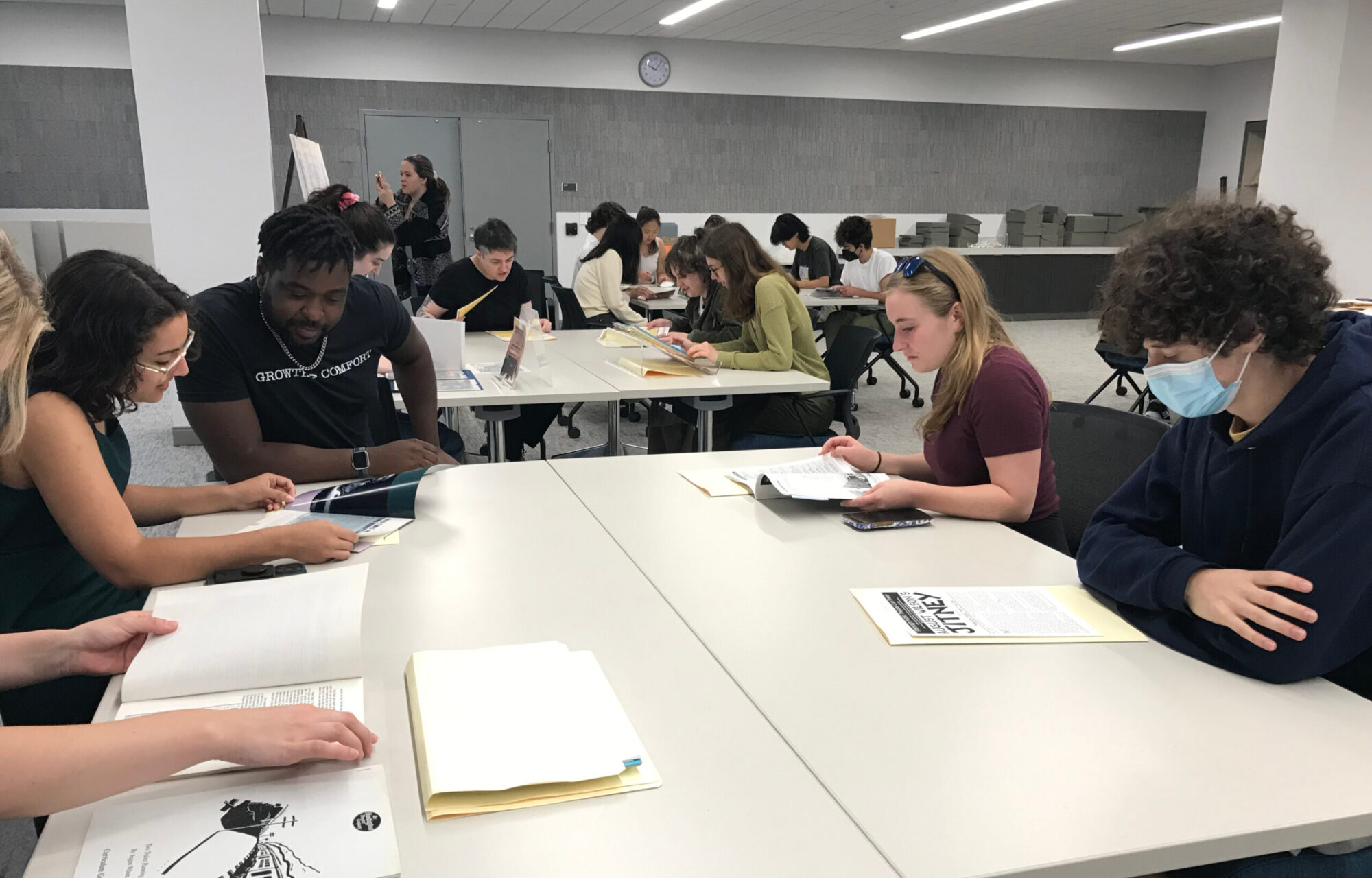 Students sit around tables, reading from books and documents.