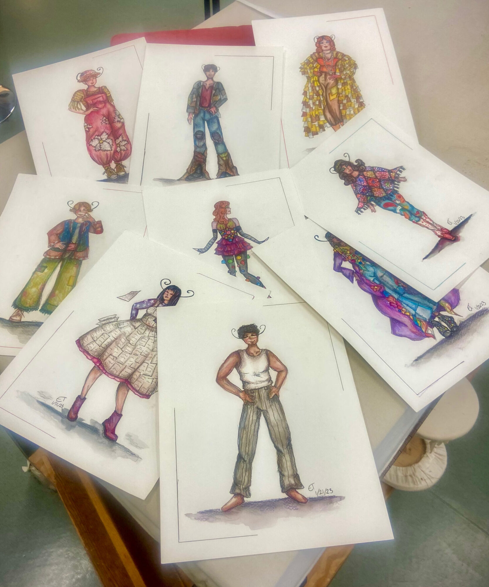 Nine colorful costume design sketches for a production of “Godspell” are spread out across a table.