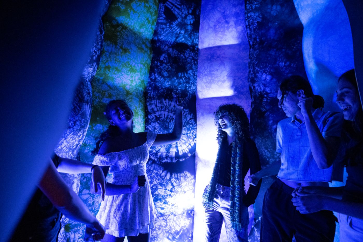 Four students laughing and dancing, surrounded by fabric in dim purple lighting.