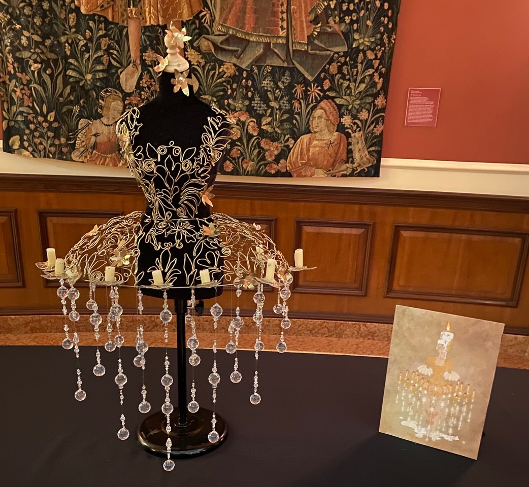 A small dress form with an intricate wire and bead gown next to a photo of the inspiration for the design - a chandelier candelabra.