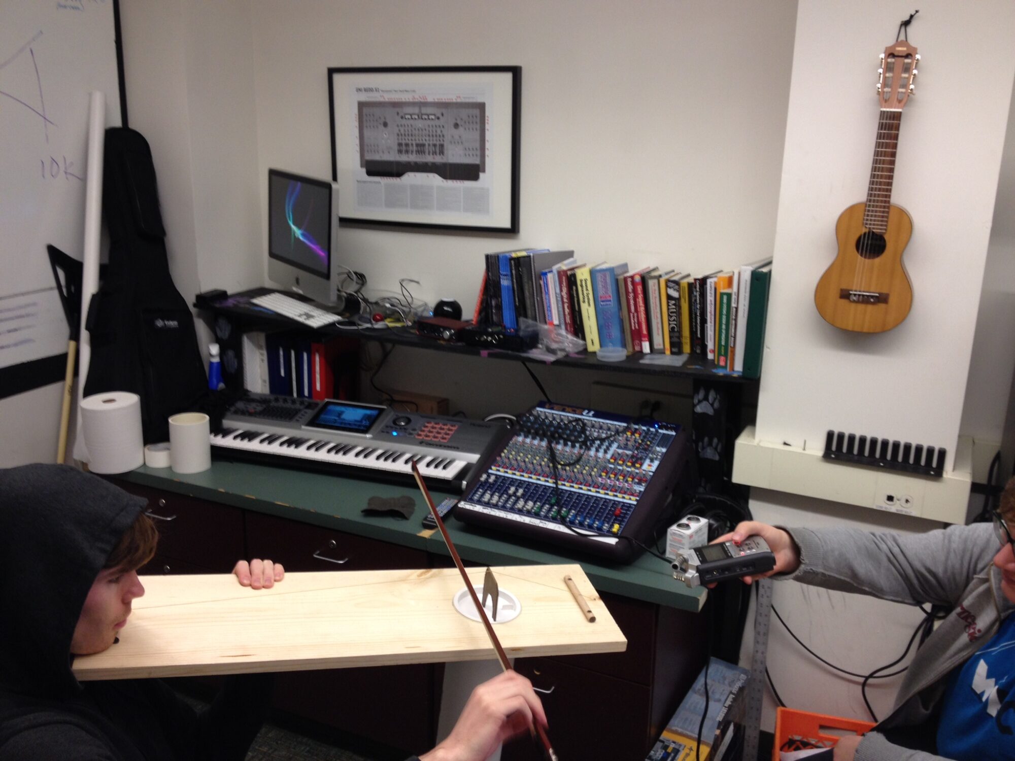 Two students in a classroom, one with a long board under his chin and a sting pulled taut over a small vertical piece of cardboard-like material; he runs a bow over the string; musical instruments and equipment in the background.