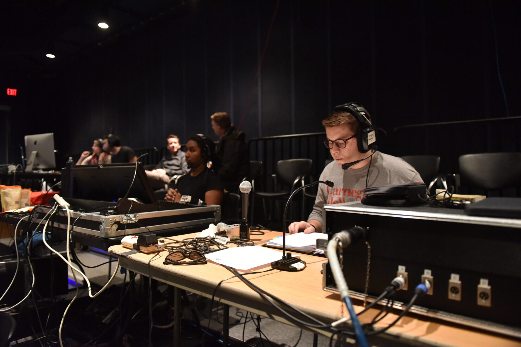 A student stage manager wears a headset and sits at a tech table with his production book open, and a microphone in front of him.