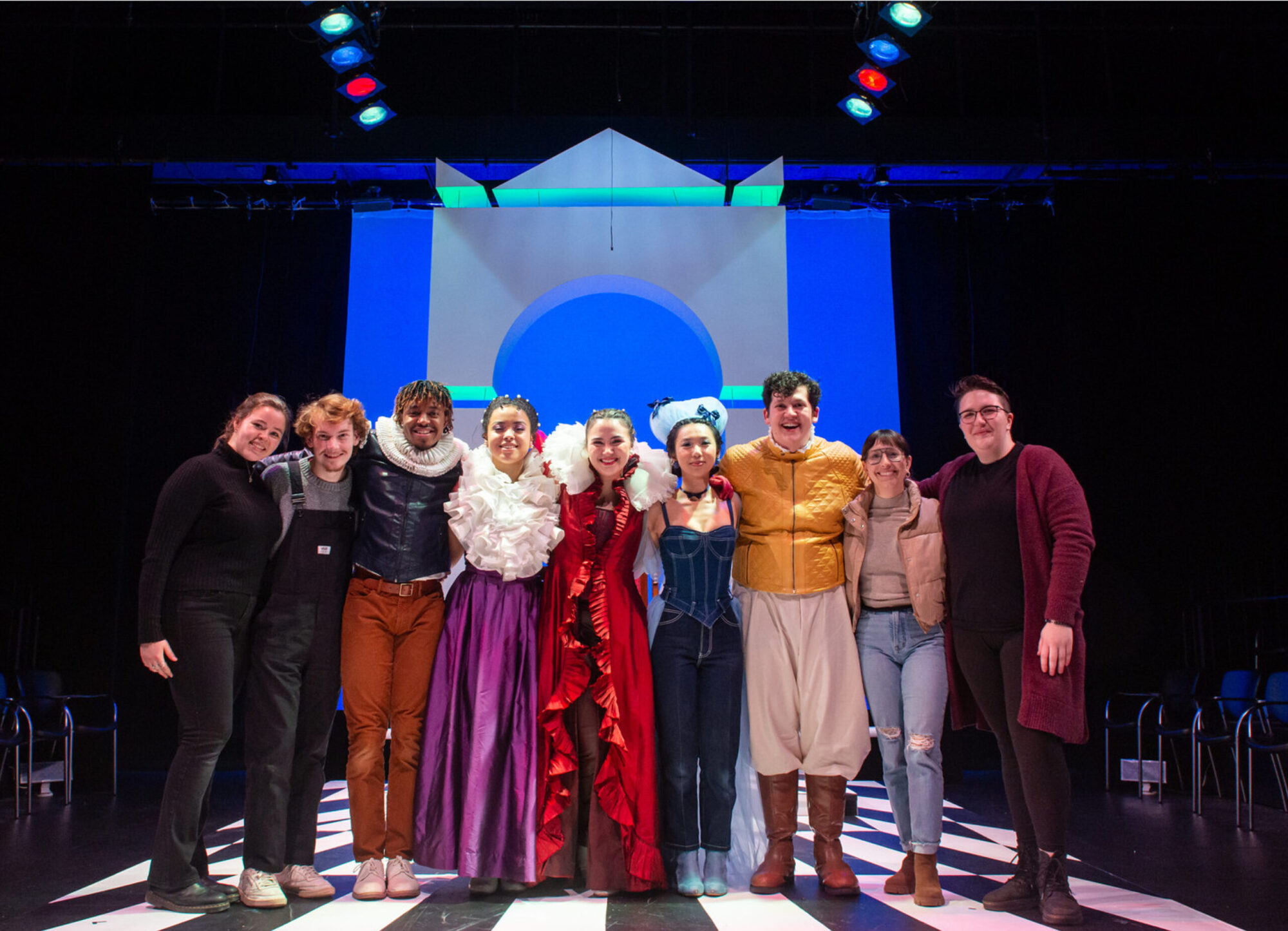 A group of students stands on the set of a play, with the arms around each other, some in costume, some wearing all black.