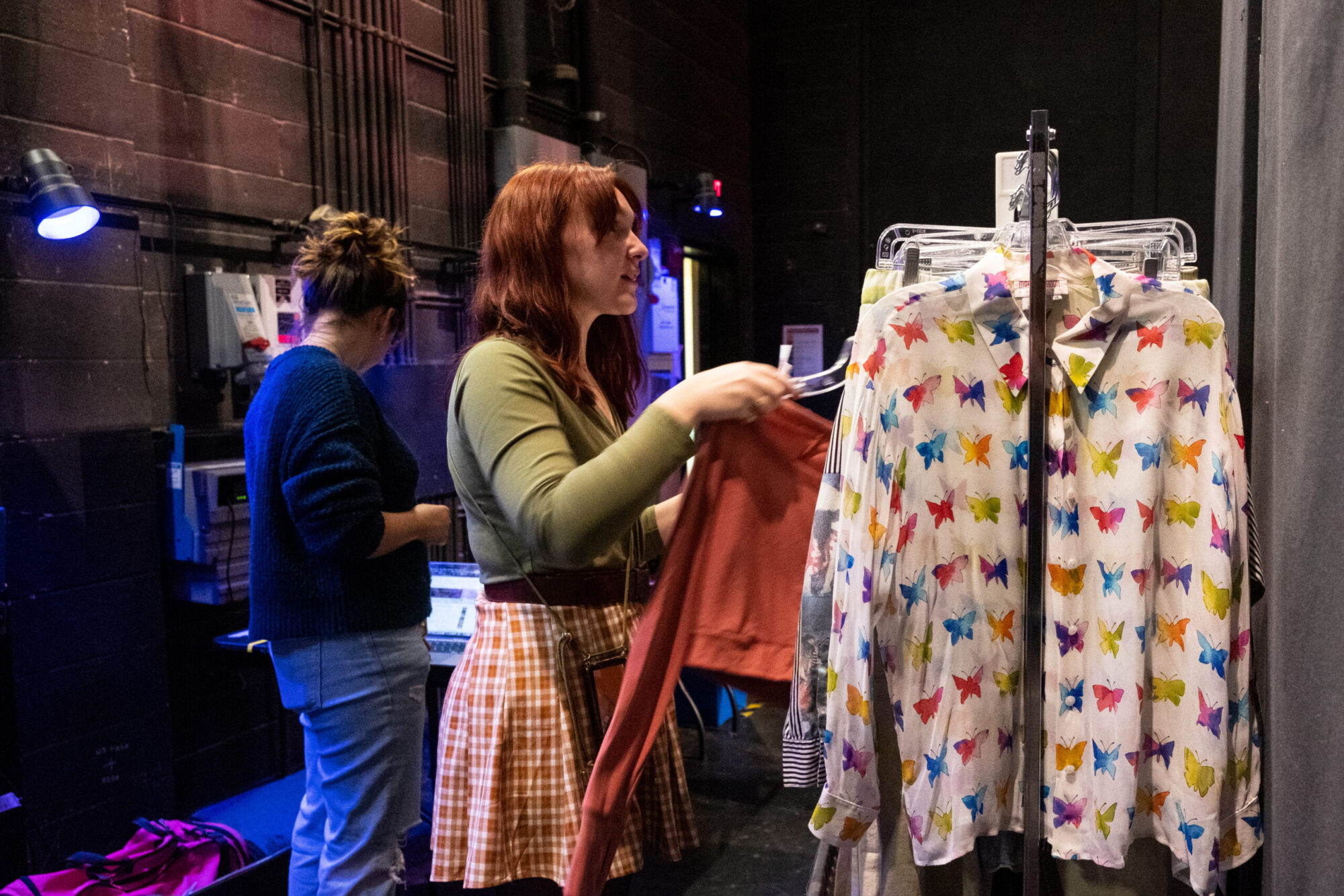 Two students are backstage in a theater, hanging costumes on a costume rack.