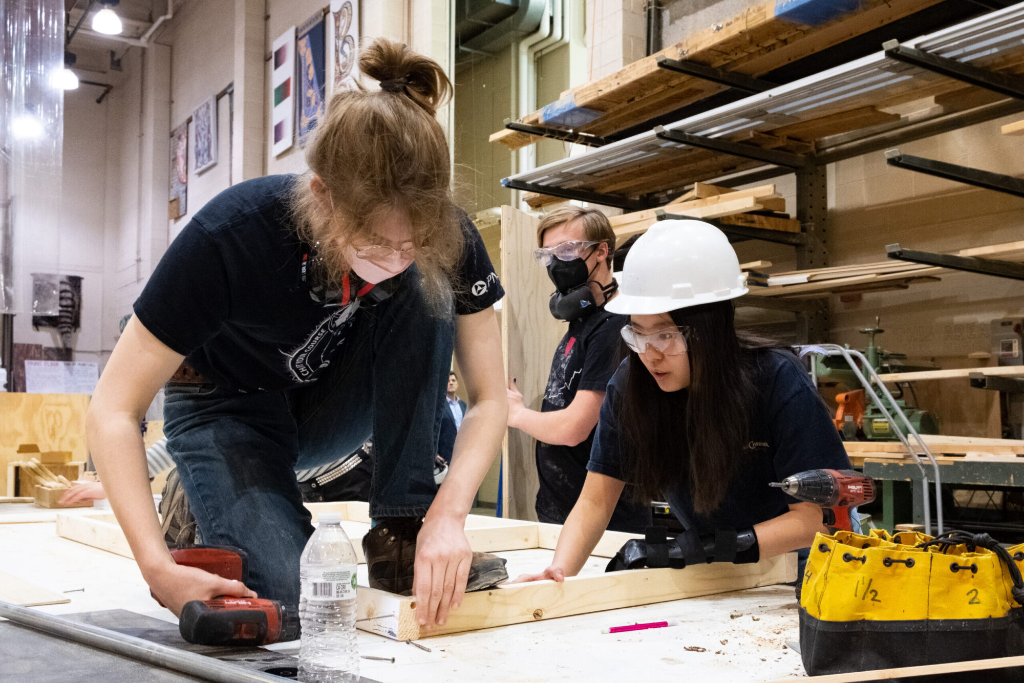 Students working in the scene shop, using a drill, wearing safety glasses and hard hats.