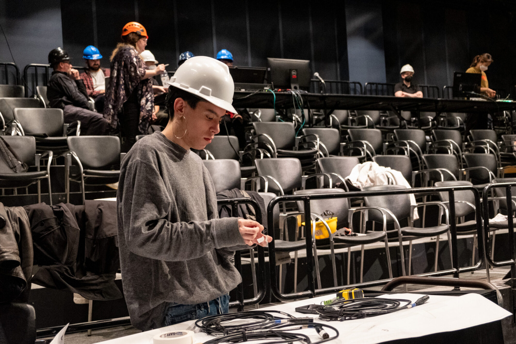 A student wearing a gray long-sleeved shirt and a white hard hat stands at a table in a black box theater, preparing cables, during a production load-in.