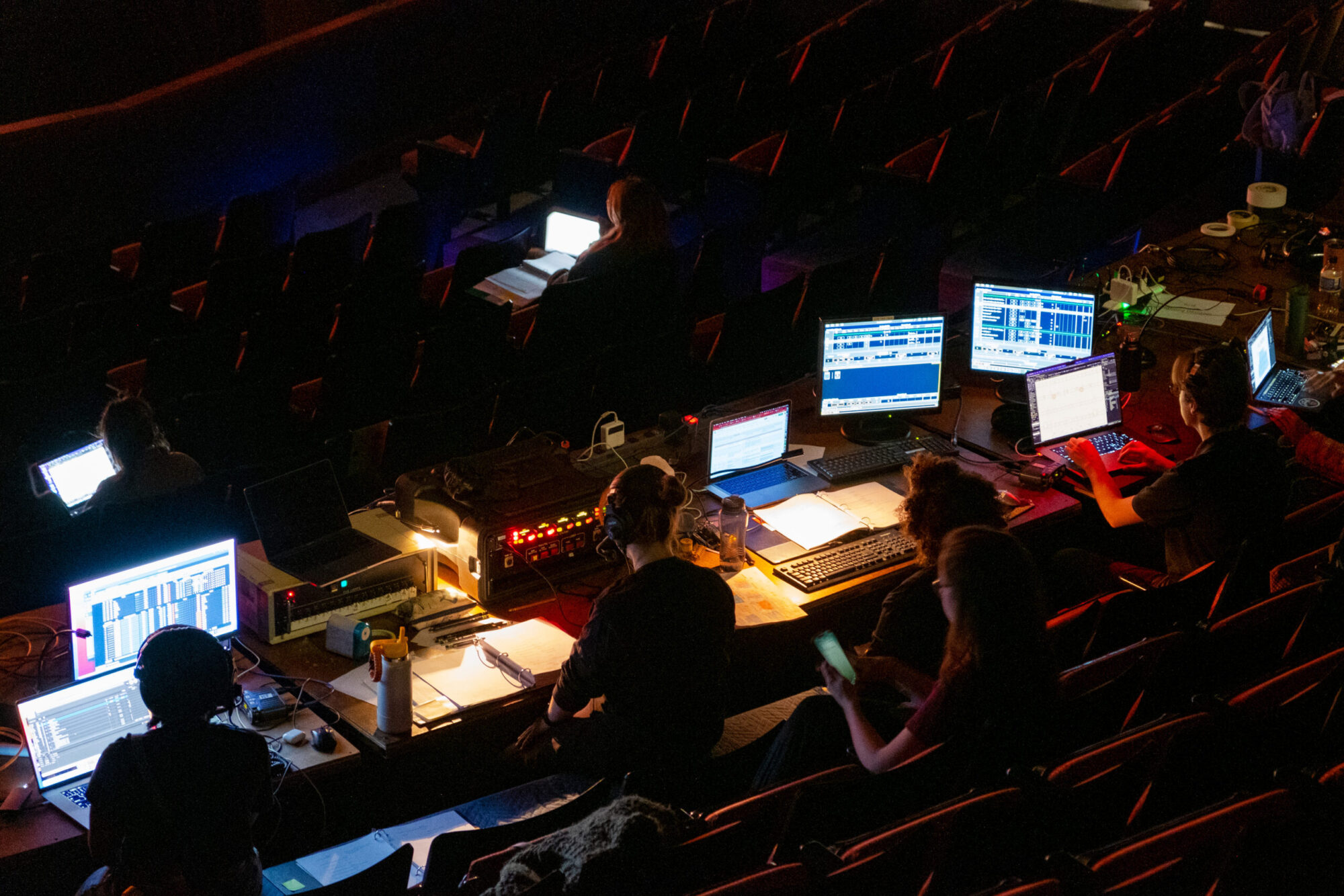 In a dark theater, a table with lit up computer screens and students sitting and working with headsets.