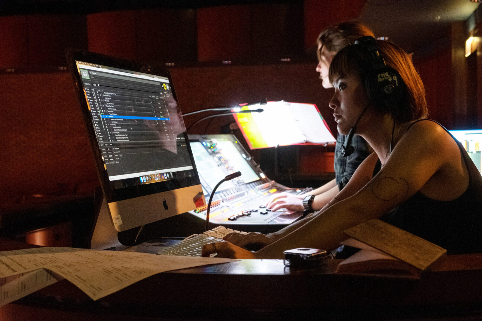 A student wearing a headset sits at a table in a darkened theater working on a large computer screen during a technical rehearsal.