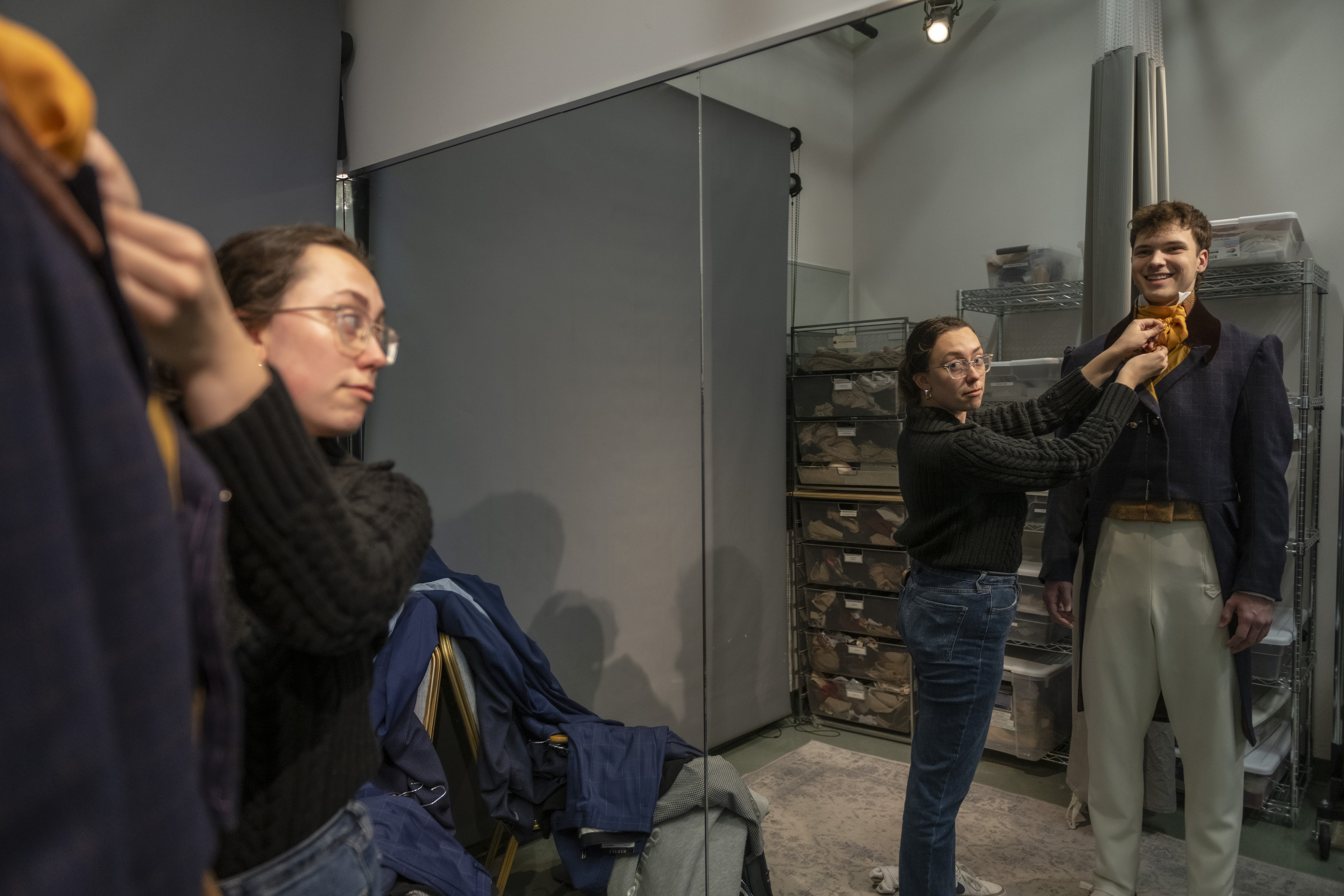 One student wears a costume, looking in a full-length mirror, while another student adjusts a scarf around his neck in a costume fitting.