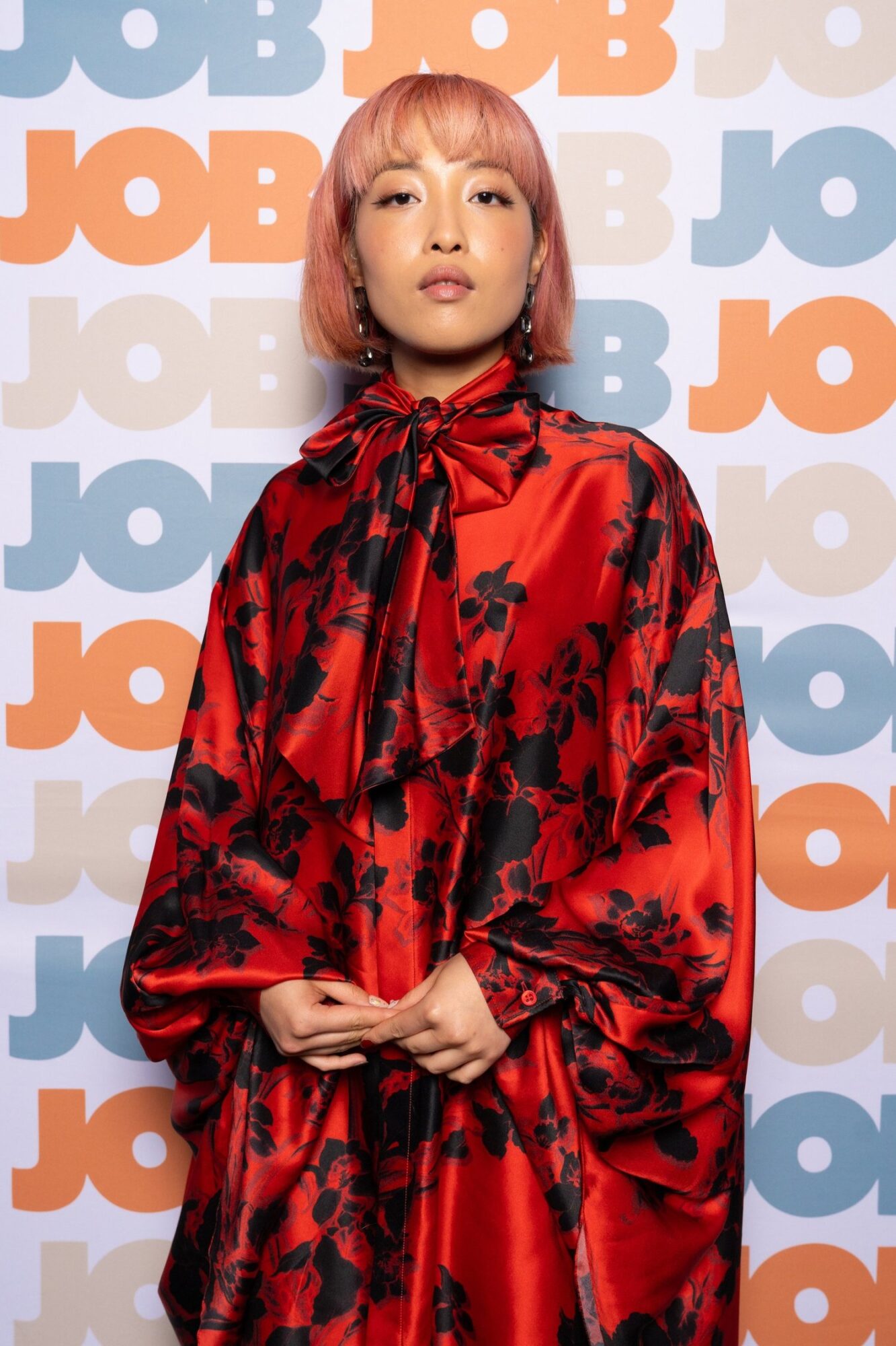 Michelle J. Li, wearing a red silk dress with black floral print and a large bow at her neck, stands in front of step and repeat that has “JOB” repeated in bold blue and orange font.