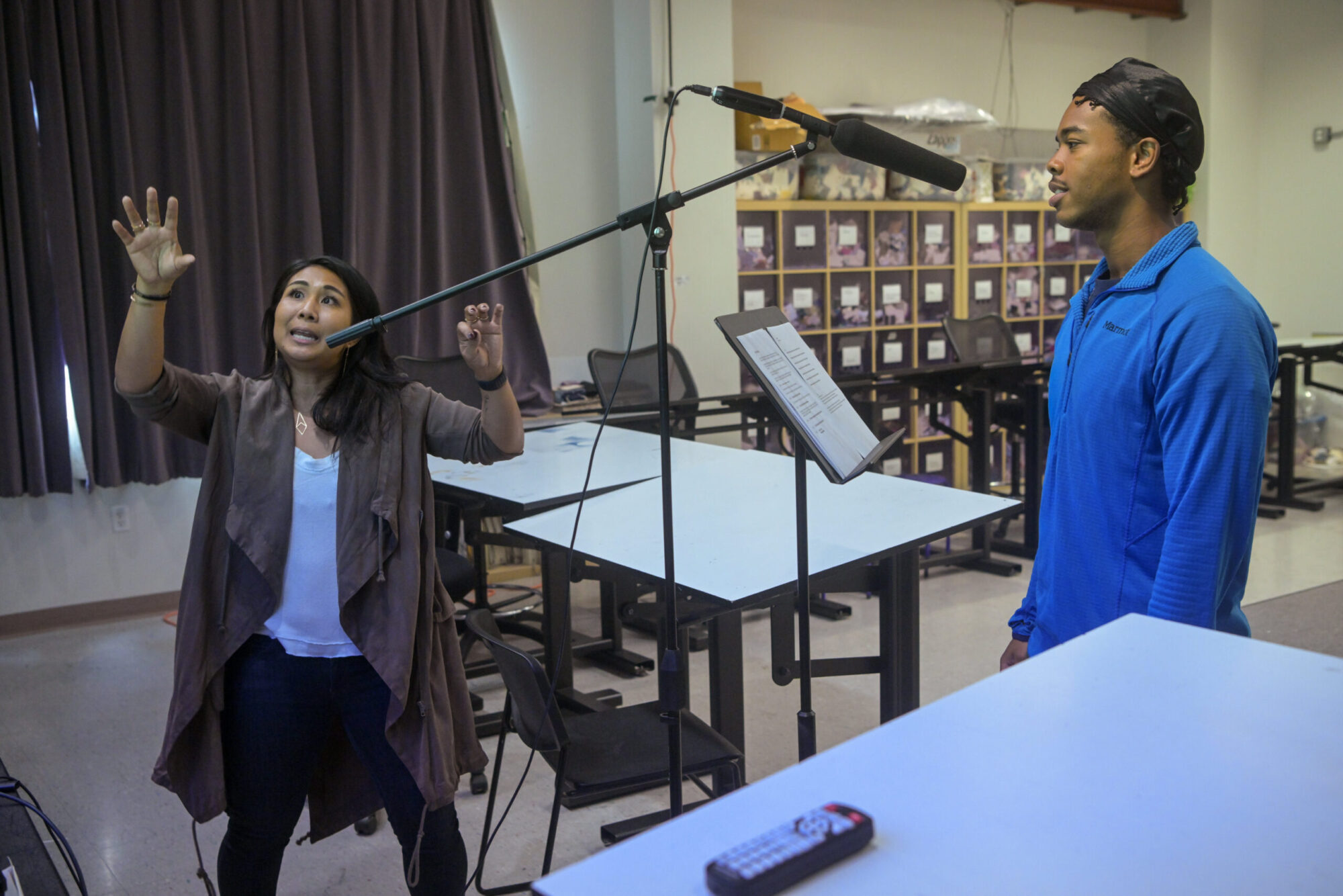 A woman wearing a long brown cardigan gestures, looking up, while a student wearing a blue shirt and black head wrap stands at a microphone in a classroom.