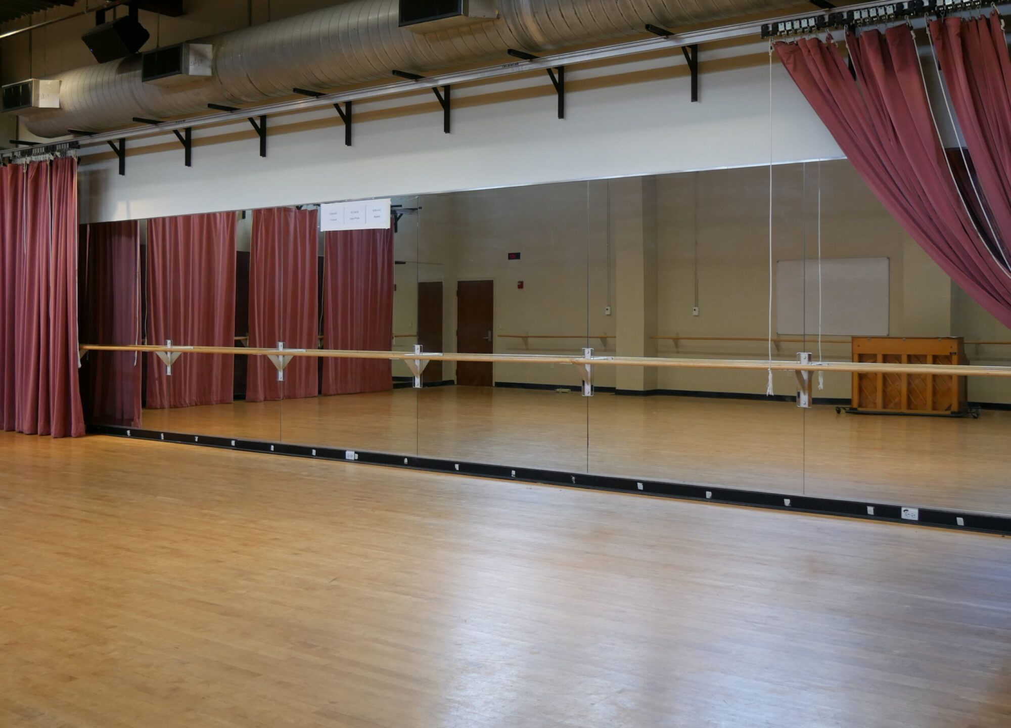 A photo of the Morie Dance Studio, with light wood floors, a wall of mirrors, a ballet bar, and red curtains hanging on the sides.