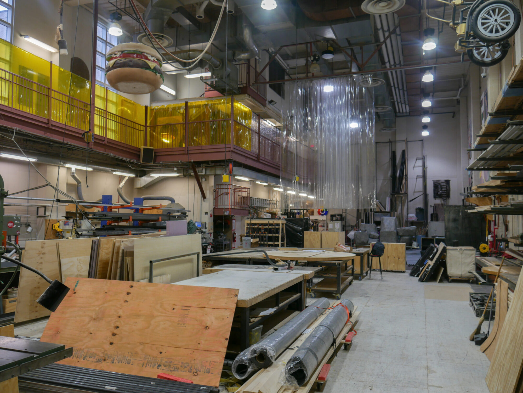 Interior photo of the large Carnegie Mellon scene shop, with various building materials and equipment, and past set pieces suspended from the ceiling.