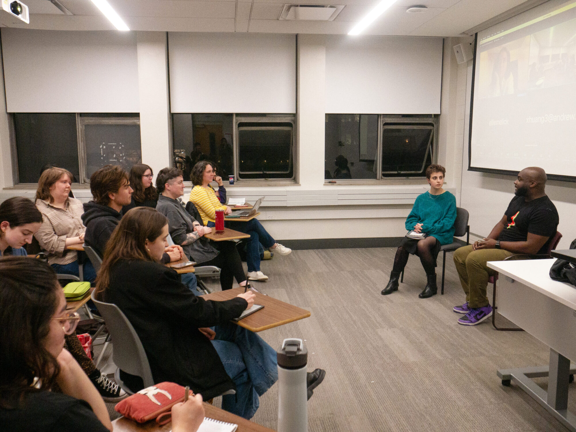 In a classroom, several students and faculty sit at desks, listening to a discussion with a playwright and student facilitator, who sit at the front of the room.