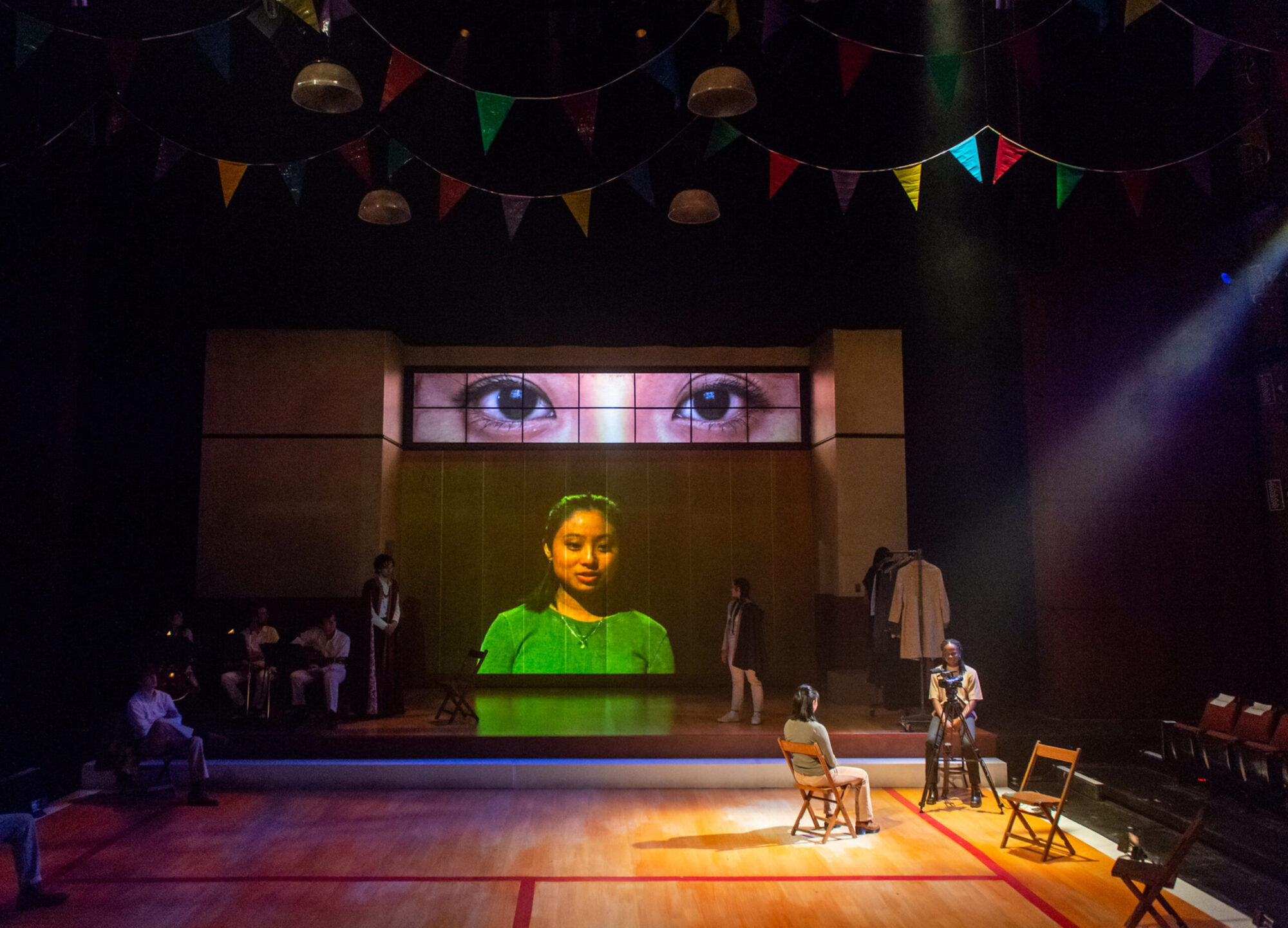 A large stage with a gymnasium floor and colorful party flags strung from the ceiling; two actors sit on stage acting each other, one actor is projected large on the back wall, with an even larger projection of just her eyes above that.