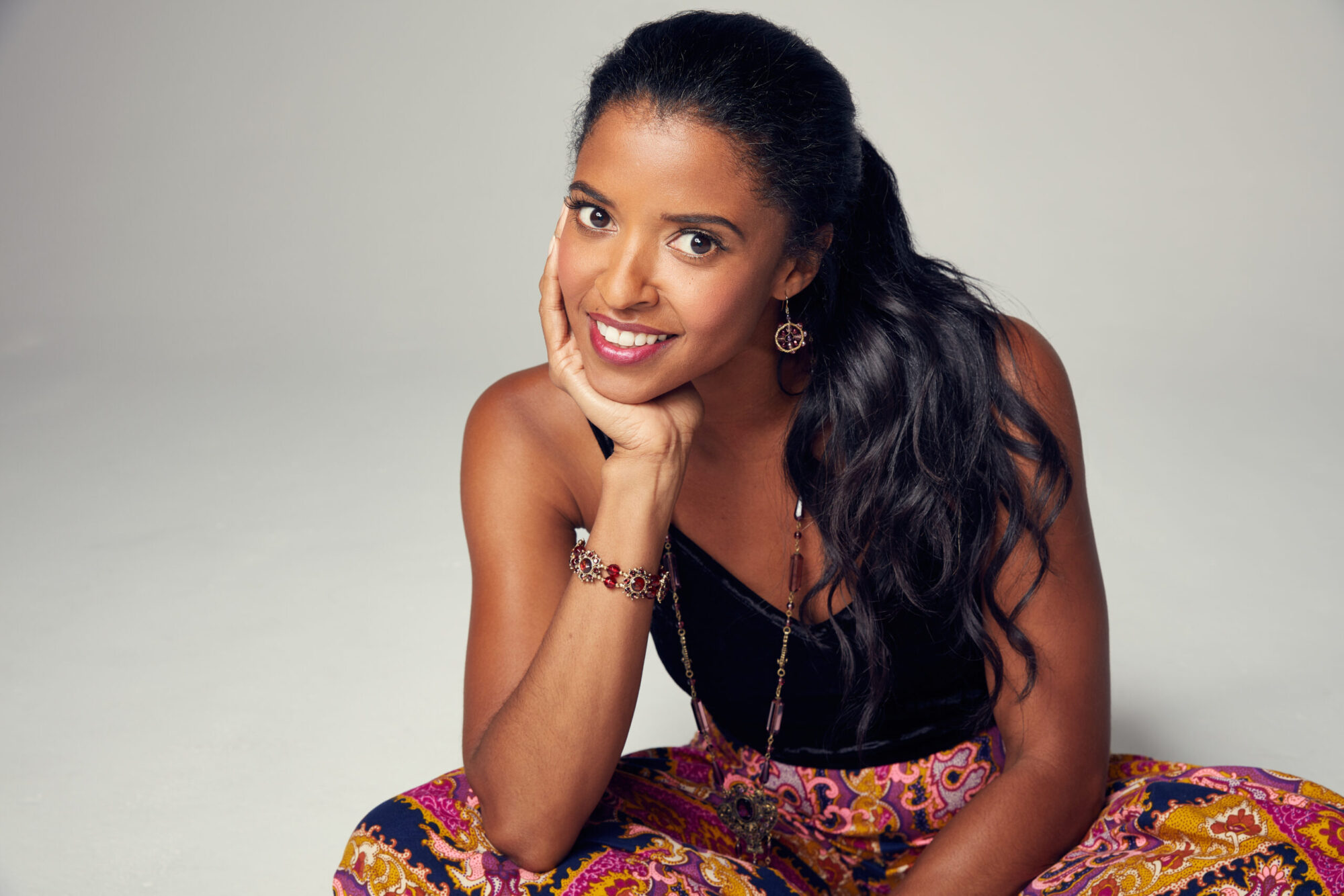 Renée Elise Goldsberry sits cross-legged, resting her chin in her hand and smiling at the camera.