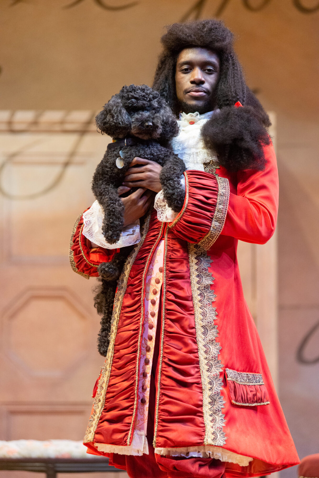 An actor wearing an elaborate red Victorian coat and wig holds small black poodle and looks directly at the camera.